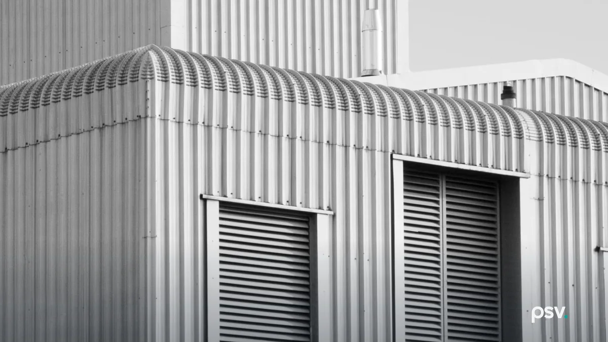 Black and white photograph of a single-story industrial building shot from below against a bright sky, its walls and barrel-vaulted roof clad in vertical corrugated metal, with two tall louvred vents set into the near elevation and a metal flue rising from a taller structure behind it