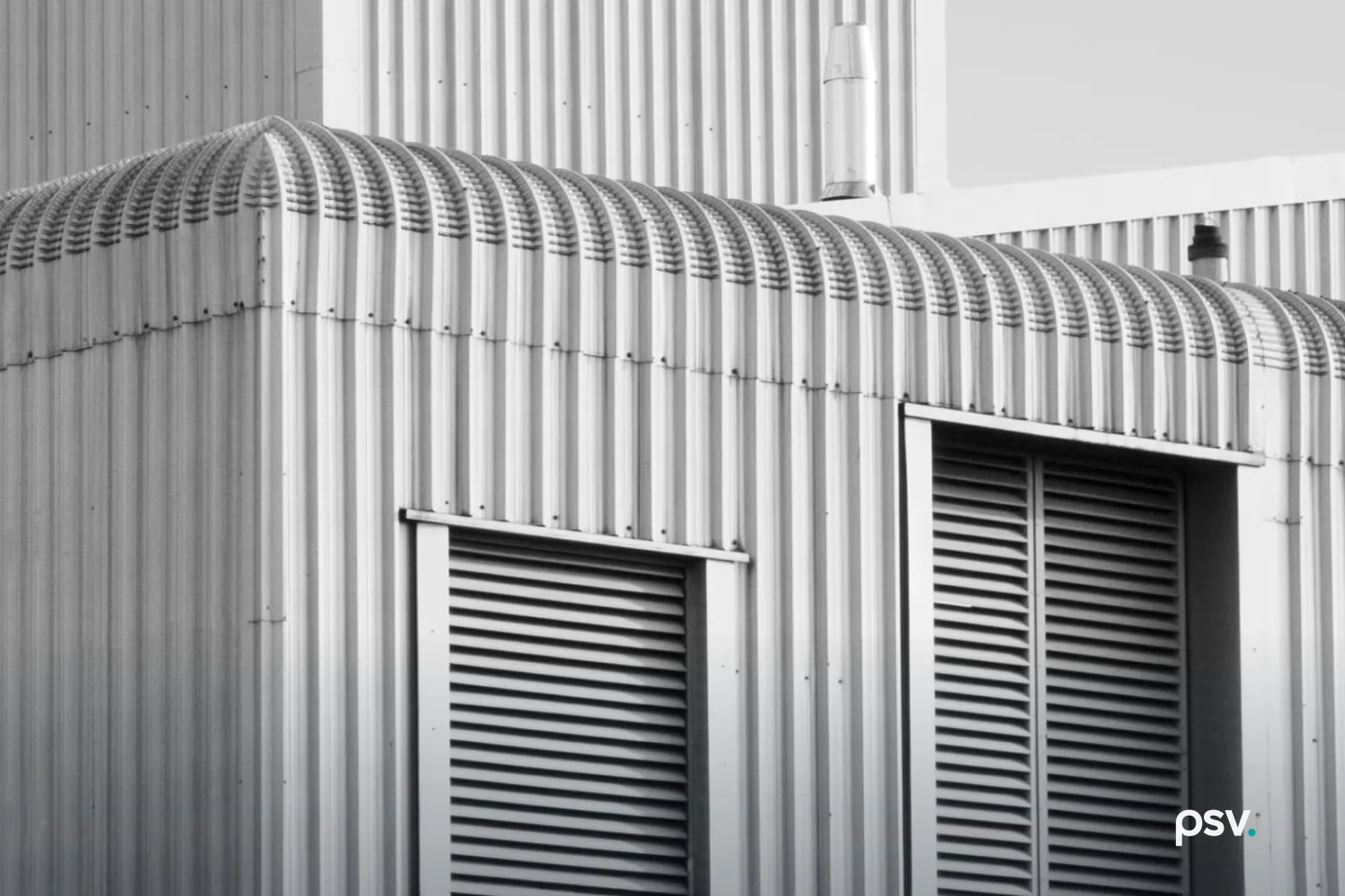 Black and white photograph of a single-story industrial building shot from below against a bright sky, its walls and barrel-vaulted roof clad in vertical corrugated metal, with two tall louvred vents set into the near elevation and a metal flue rising from a taller structure behind it Black and white photograph of a single-story industrial building shot from below against a bright sky, its walls and barrel-vaulted roof clad in vertical corrugated metal, with two tall louvred vents set into the near elevation and a metal flue rising from a taller structure behind it