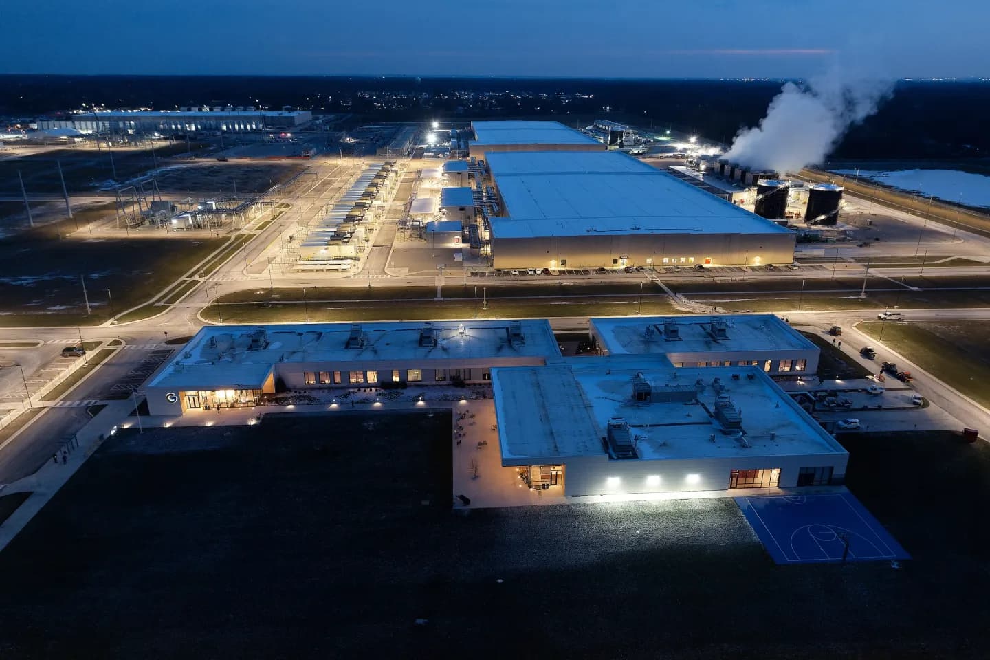 Aerial view at dusk of Google's New Albany, Ohio data center campus: a large illuminated server hall beside an electrical substation yard, with operations offices lit in the foreground Aerial view at dusk of Google's New Albany, Ohio data center campus: a large illuminated server hall beside an electrical substation yard, with operations offices lit in the foreground