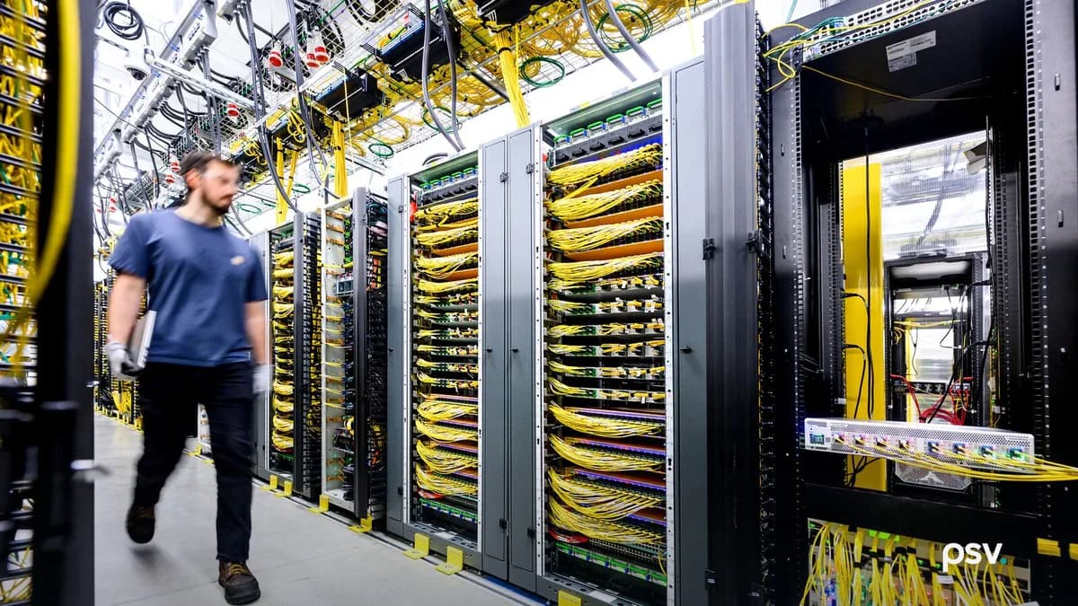 The inside of a brightly lit data center, with a long row of open server and network racks filled with bundled yellow fiber optic cables, overhead cable trays running along the ceiling, and a technician in a blue T-shirt and work gloves walking down the aisle at the left carrying a laptop.