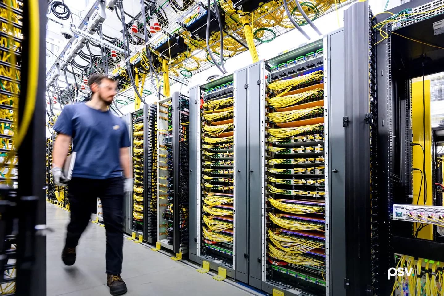 The inside of a brightly lit data center, with a long row of open server and network racks filled with bundled yellow fiber optic cables, overhead cable trays running along the ceiling, and a technician in a blue T-shirt and work gloves walking down the aisle at the left carrying a laptop. The inside of a brightly lit data center, with a long row of open server and network racks filled with bundled yellow fiber optic cables, overhead cable trays running along the ceiling, and a technician in a blue T-shirt and work gloves walking down the aisle at the left carrying a laptop.