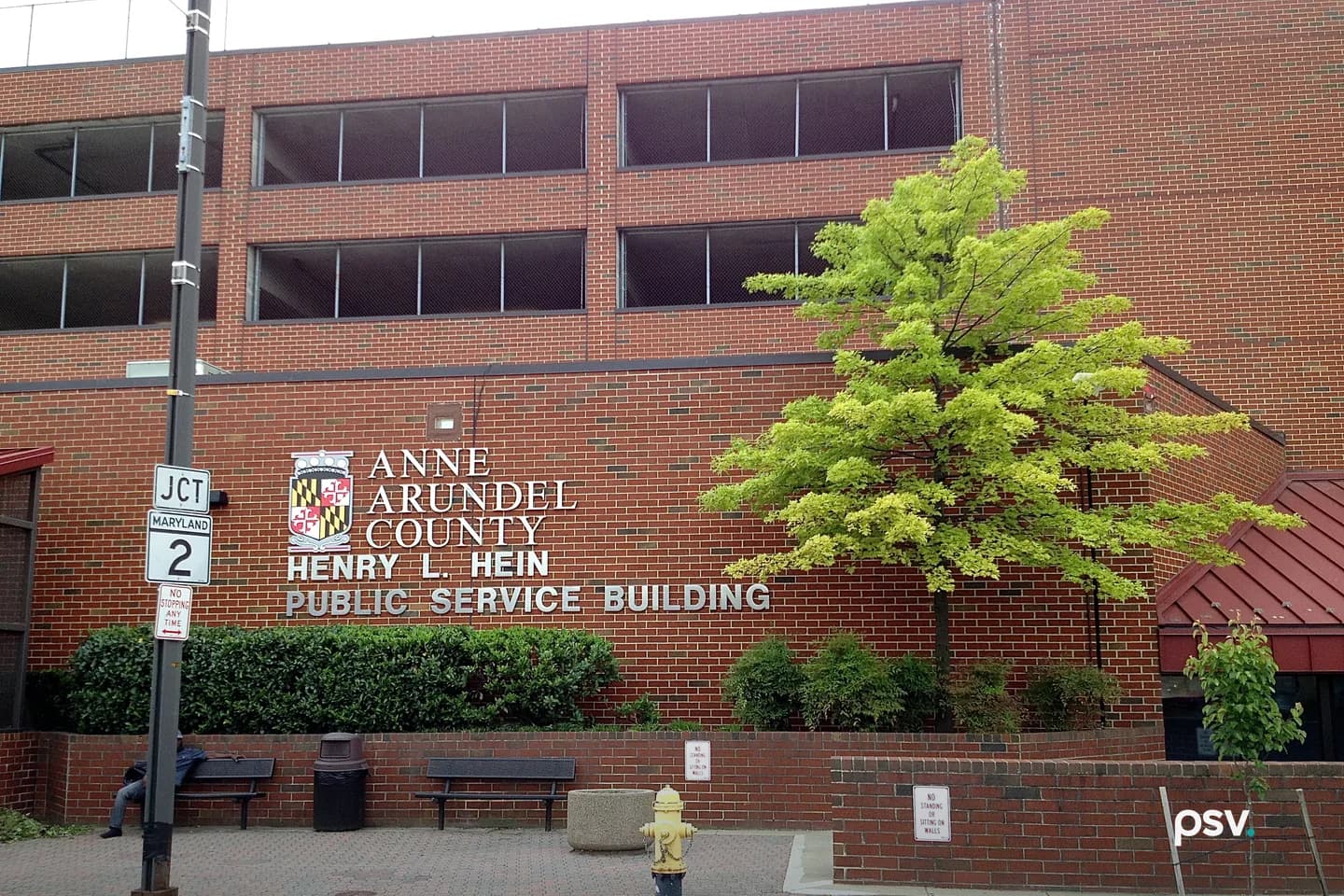 A red brick building with rows of dark horizontal openings on its upper floors, metal lettering on the wall reading Anne Arundel County, Henry L. Hein Public Service Building beside the county seal, a trimmed hedge and two benches along a brick sidewalk, a young tree with bright green leaves at the right, a yellow fire hydrant at the curb and a Maryland Route 2 junction sign on a pole at the left. A red brick building with rows of dark horizontal openings on its upper floors, metal lettering on the wall reading Anne Arundel County, Henry L. Hein Public Service Building beside the county seal, a trimmed hedge and two benches along a brick sidewalk, a young tree with bright green leaves at the right, a yellow fire hydrant at the curb and a Maryland Route 2 junction sign on a pole at the left.