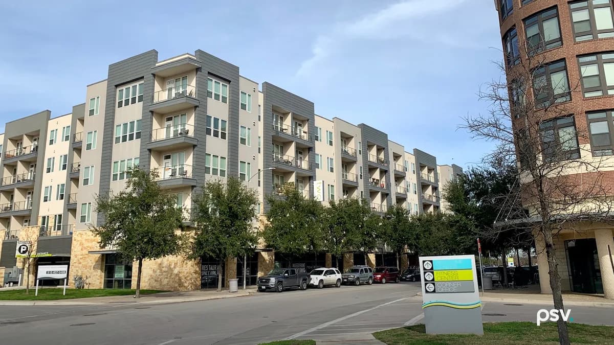 A daylight photograph of a five-storey apartment building in Austin, Texas, clad in grey and cream panels with stacked balconies, a stone-faced ground floor of shopfronts behind a row of young live oaks, pickup trucks and cars parked along the street, a rounded brick corner building with tall windows at the right edge, and a wayfinding sign in the foreground with arrows pointing to a fitness center and a corner office, under a blue sky with thin cloud.