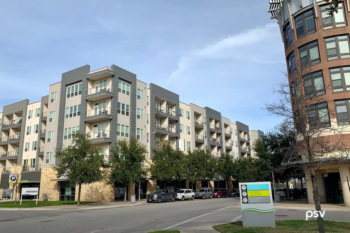 A daylight photograph of a five-storey apartment building in Austin, Texas, clad in grey and cream panels with stacked balconies, a stone-faced ground floor of shopfronts behind a row of young live oaks, pickup trucks and cars parked along the street, a rounded brick corner building with tall windows at the right edge, and a wayfinding sign in the foreground with arrows pointing to a fitness center and a corner office, under a blue sky with thin cloud. A daylight photograph of a five-storey apartment building in Austin, Texas, clad in grey and cream panels with stacked balconies, a stone-faced ground floor of shopfronts behind a row of young live oaks, pickup trucks and cars parked along the street, a rounded brick corner building with tall windows at the right edge, and a wayfinding sign in the foreground with arrows pointing to a fitness center and a corner office, under a blue sky with thin cloud.