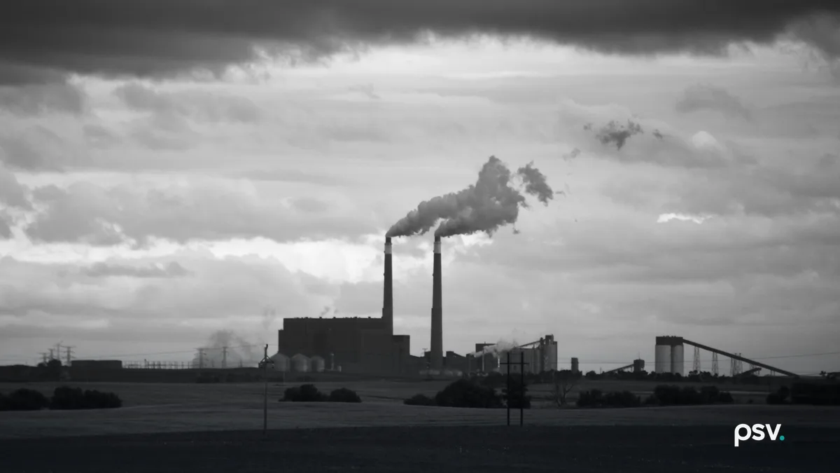 A power station on a flat horizon seen across dark farm fields, with two tall smokestacks releasing plumes that drift to the right, a large boiler building at their base, silos and an inclined conveyor to the right, transmission towers at the far left, two wooden utility poles in the foreground and a heavy bank of cloud across the top of the sky.