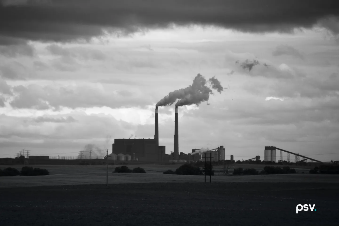 A power station on a flat horizon seen across dark farm fields, with two tall smokestacks releasing plumes that drift to the right, a large boiler building at their base, silos and an inclined conveyor to the right, transmission towers at the far left, two wooden utility poles in the foreground and a heavy bank of cloud across the top of the sky. A power station on a flat horizon seen across dark farm fields, with two tall smokestacks releasing plumes that drift to the right, a large boiler building at their base, silos and an inclined conveyor to the right, transmission towers at the far left, two wooden utility poles in the foreground and a heavy bank of cloud across the top of the sky.