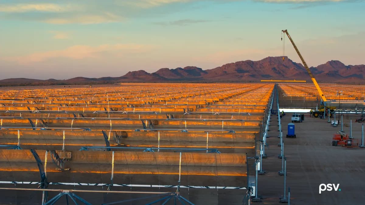 A wide golden-hour view across a utility-scale solar thermal field under construction in the Arizona desert: dozens of parallel rows of curved parabolic trough mirrors on steel frames stretching to the horizon and catching orange sunset light, a yellow mobile crane with its boom raised at right lifting a long assembly, a white pickup truck, a blue portable toilet and orange lift equipment on the graded dirt lane between the rows, and a low red rock mountain ridge across the background under a pale blue and pink sky.