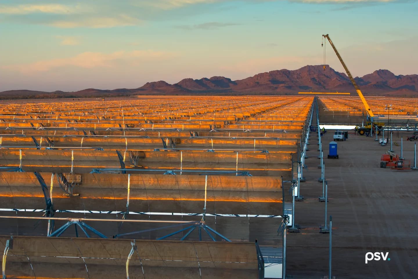 A wide golden-hour view across a utility-scale solar thermal field under construction in the Arizona desert: dozens of parallel rows of curved parabolic trough mirrors on steel frames stretching to the horizon and catching orange sunset light, a yellow mobile crane with its boom raised at right lifting a long assembly, a white pickup truck, a blue portable toilet and orange lift equipment on the graded dirt lane between the rows, and a low red rock mountain ridge across the background under a pale blue and pink sky. A wide golden-hour view across a utility-scale solar thermal field under construction in the Arizona desert: dozens of parallel rows of curved parabolic trough mirrors on steel frames stretching to the horizon and catching orange sunset light, a yellow mobile crane with its boom raised at right lifting a long assembly, a white pickup truck, a blue portable toilet and orange lift equipment on the graded dirt lane between the rows, and a low red rock mountain ridge across the background under a pale blue and pink sky.