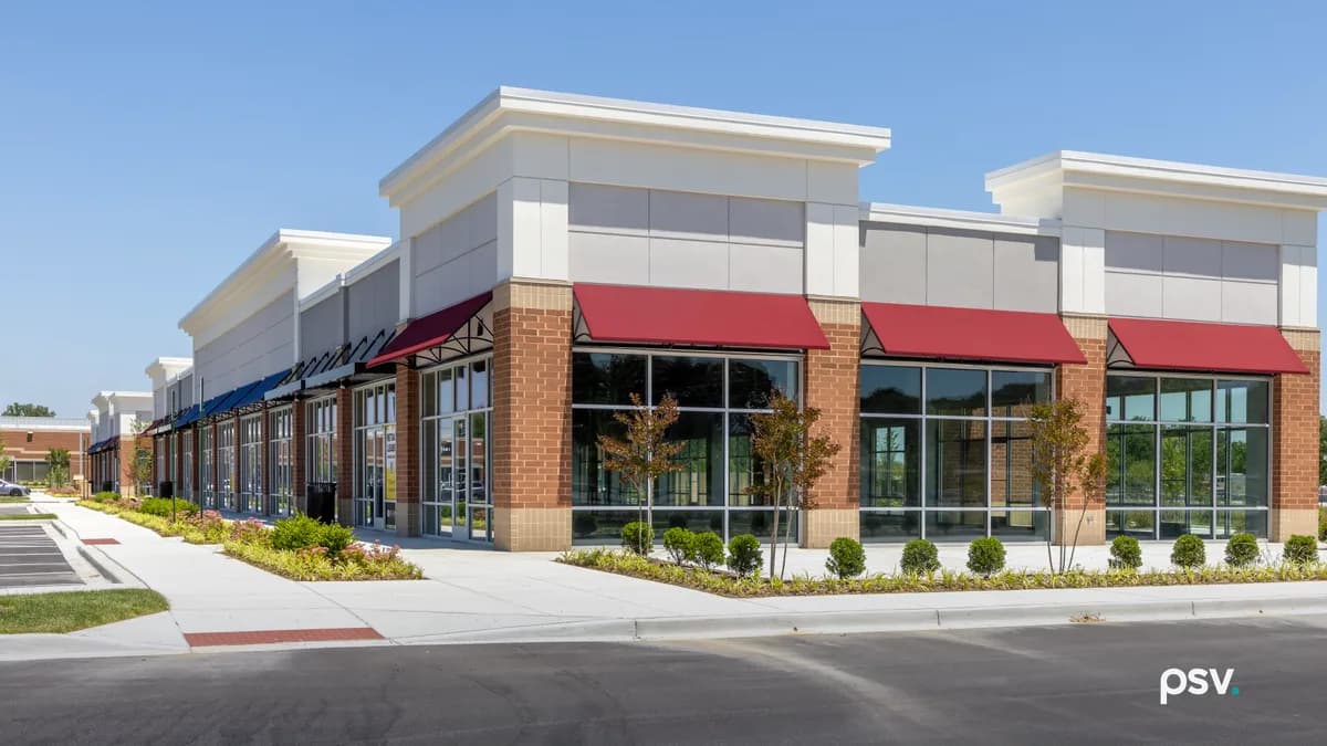 A daylight photograph of a newly built single-storey multi-tenant retail building with red brick piers, cream parapets, tall glass shopfronts and red and blue fabric awnings, its units still empty, fronted by a new concrete sidewalk, young ornamental trees and flowerbeds, with a freshly paved parking lot in the foreground and a clear blue sky above.