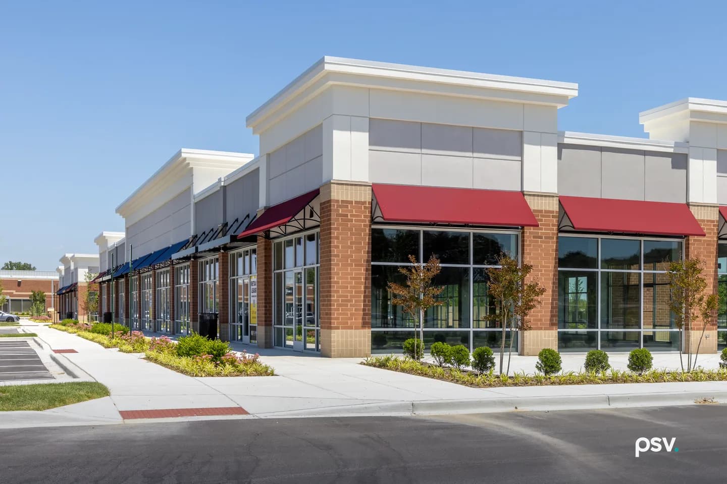A daylight photograph of a newly built single-storey multi-tenant retail building with red brick piers, cream parapets, tall glass shopfronts and red and blue fabric awnings, its units still empty, fronted by a new concrete sidewalk, young ornamental trees and flowerbeds, with a freshly paved parking lot in the foreground and a clear blue sky above. A daylight photograph of a newly built single-storey multi-tenant retail building with red brick piers, cream parapets, tall glass shopfronts and red and blue fabric awnings, its units still empty, fronted by a new concrete sidewalk, young ornamental trees and flowerbeds, with a freshly paved parking lot in the foreground and a clear blue sky above.