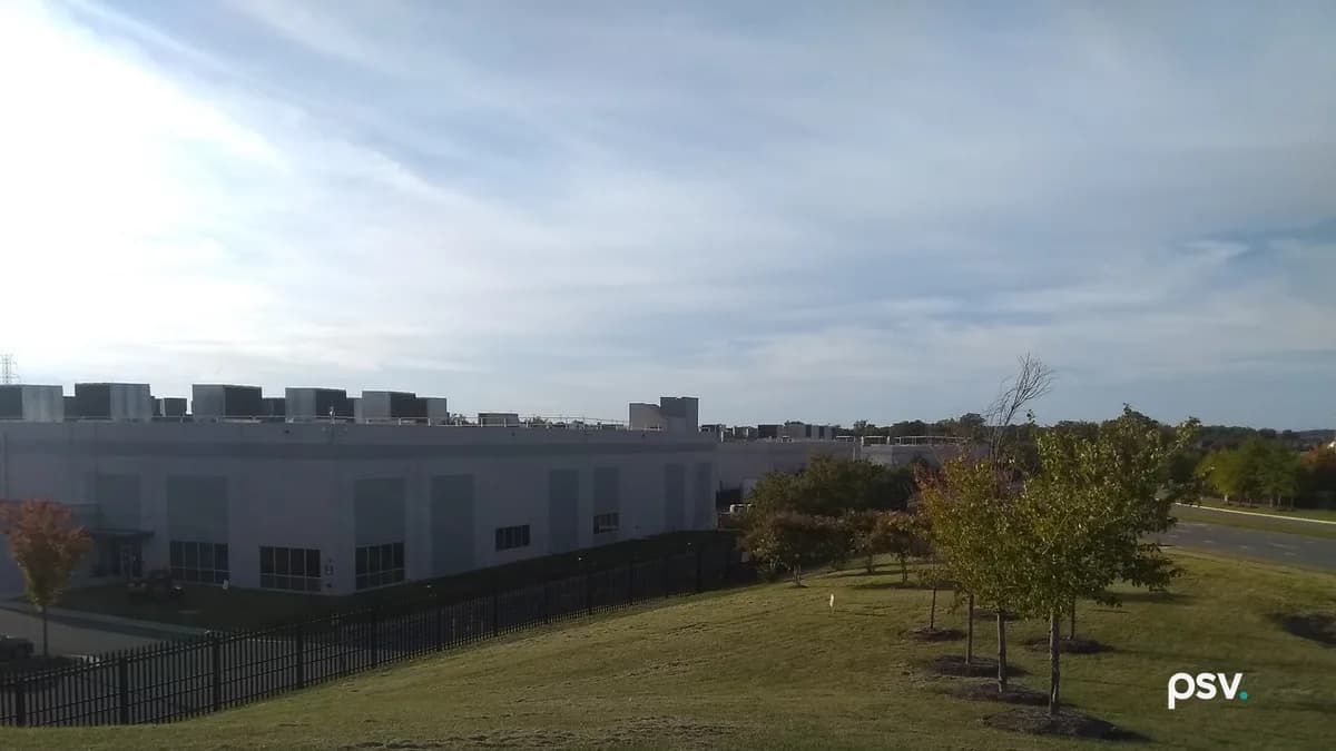 A long, low grey data center building with rows of dark rooftop cooling units stands behind a black steel picket fence at the foot of a mown grass slope, with a line of young trees in early autumn color running down the slope at the right, a road beyond them, and a bright sky streaked with thin cloud.