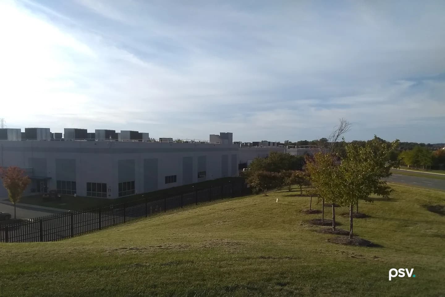A long, low grey data center building with rows of dark rooftop cooling units stands behind a black steel picket fence at the foot of a mown grass slope, with a line of young trees in early autumn color running down the slope at the right, a road beyond them, and a bright sky streaked with thin cloud. A long, low grey data center building with rows of dark rooftop cooling units stands behind a black steel picket fence at the foot of a mown grass slope, with a line of young trees in early autumn color running down the slope at the right, a road beyond them, and a bright sky streaked with thin cloud.
