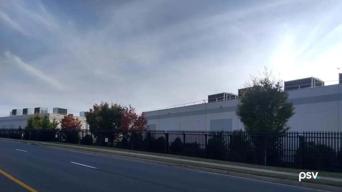 A long, windowless pale grey data center building seen from across a four-lane road, behind a black steel picket fence and a row of shrubs and trees, some with red autumn leaves, with boxy mechanical units and railings along the roofline and a second building with rooftop units further left, under a pale sky streaked with thin cloud.