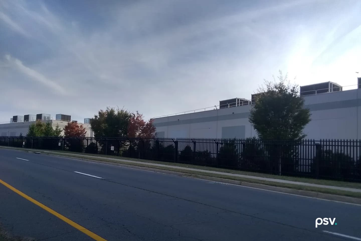 A long, windowless pale grey data center building seen from across a four-lane road, behind a black steel picket fence and a row of shrubs and trees, some with red autumn leaves, with boxy mechanical units and railings along the roofline and a second building with rooftop units further left, under a pale sky streaked with thin cloud. A long, windowless pale grey data center building seen from across a four-lane road, behind a black steel picket fence and a row of shrubs and trees, some with red autumn leaves, with boxy mechanical units and railings along the roofline and a second building with rooftop units further left, under a pale sky streaked with thin cloud.