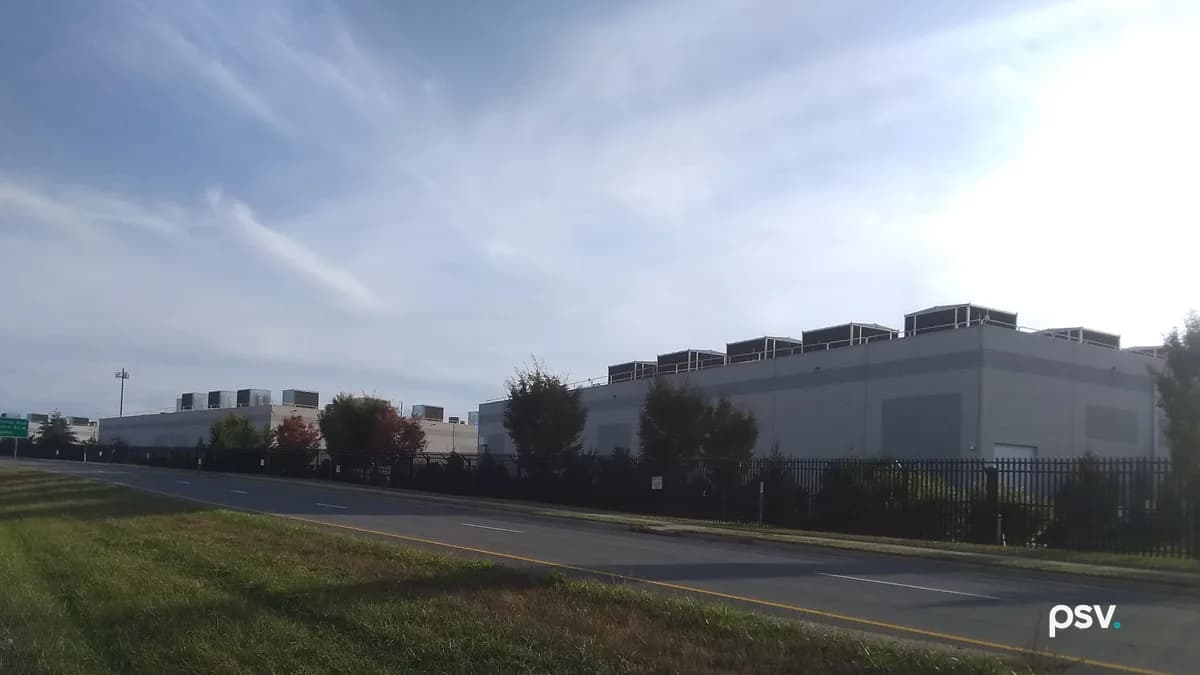 A daylight roadside photograph of large windowless grey data center buildings behind a tall black metal security fence: a row of dark rooftop equipment enclosures lines the roof of the nearest building on the right, smaller white buildings with rooftop units sit farther back on the left beside a green highway sign and a light pole, young trees stand along the fence, and a two-lane road with a yellow edge line curves across the frame in front of a mown grass verge under a pale blue sky streaked with thin clouds.