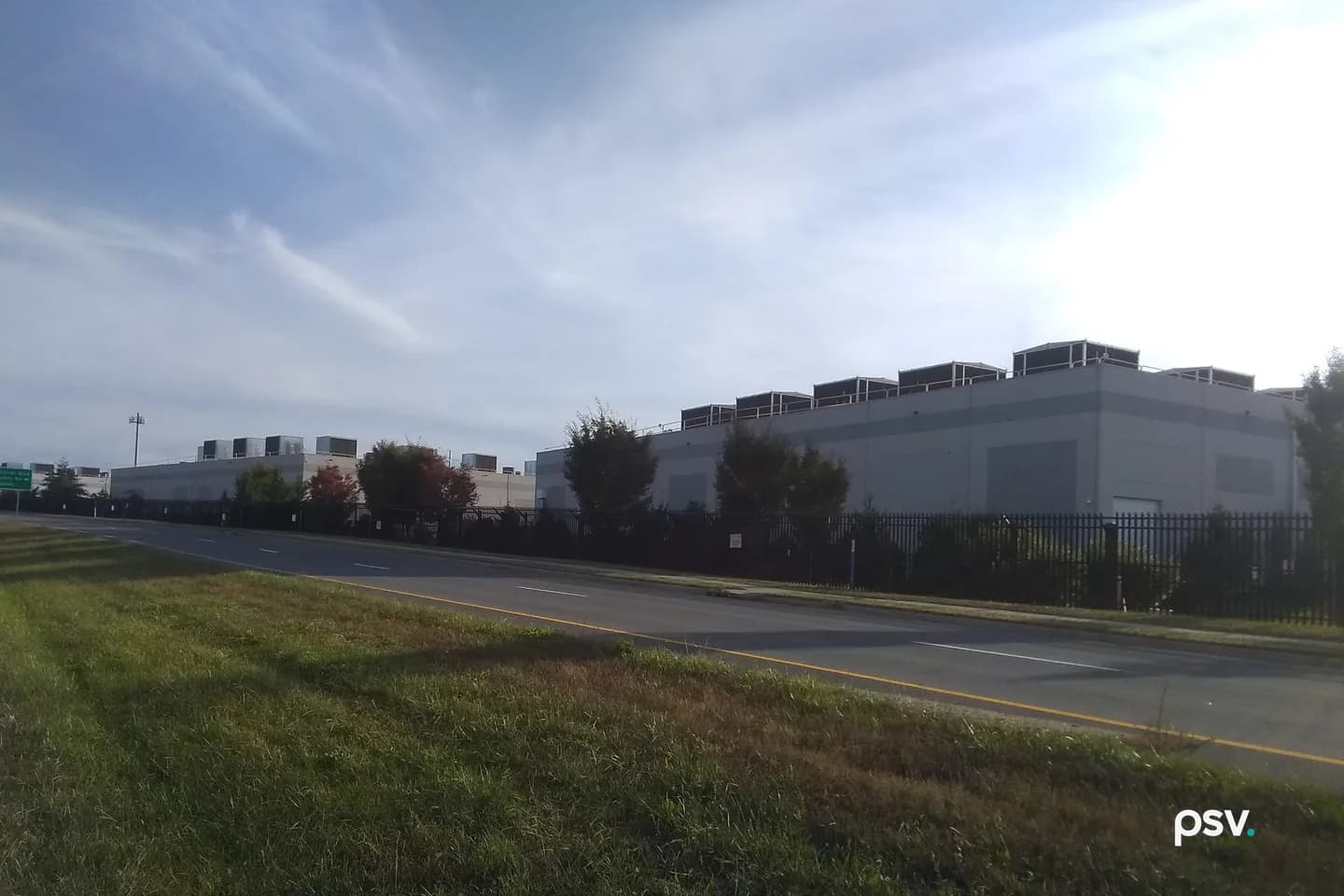 A daylight roadside photograph of large windowless grey data center buildings behind a tall black metal security fence: a row of dark rooftop equipment enclosures lines the roof of the nearest building on the right, smaller white buildings with rooftop units sit farther back on the left beside a green highway sign and a light pole, young trees stand along the fence, and a two-lane road with a yellow edge line curves across the frame in front of a mown grass verge under a pale blue sky streaked with thin clouds. A daylight roadside photograph of large windowless grey data center buildings behind a tall black metal security fence: a row of dark rooftop equipment enclosures lines the roof of the nearest building on the right, smaller white buildings with rooftop units sit farther back on the left beside a green highway sign and a light pole, young trees stand along the fence, and a two-lane road with a yellow edge line curves across the frame in front of a mown grass verge under a pale blue sky streaked with thin clouds.