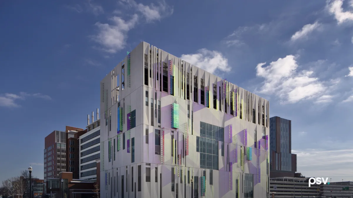 A daylight street-level view of a four-storey central chiller plant on a winter day: a boxy concrete building clad in white panels printed with pale lavender and mint geometric shapes, with narrow vertical fins along the roofline, slotted openings revealing dark mechanical equipment on the top floor, and small iridescent coloured panels scattered across the facade, with a brick hospital block to the left, a parking structure to the right, snow on the verge and a bare asphalt road in front under a blue sky with scattered cloud.