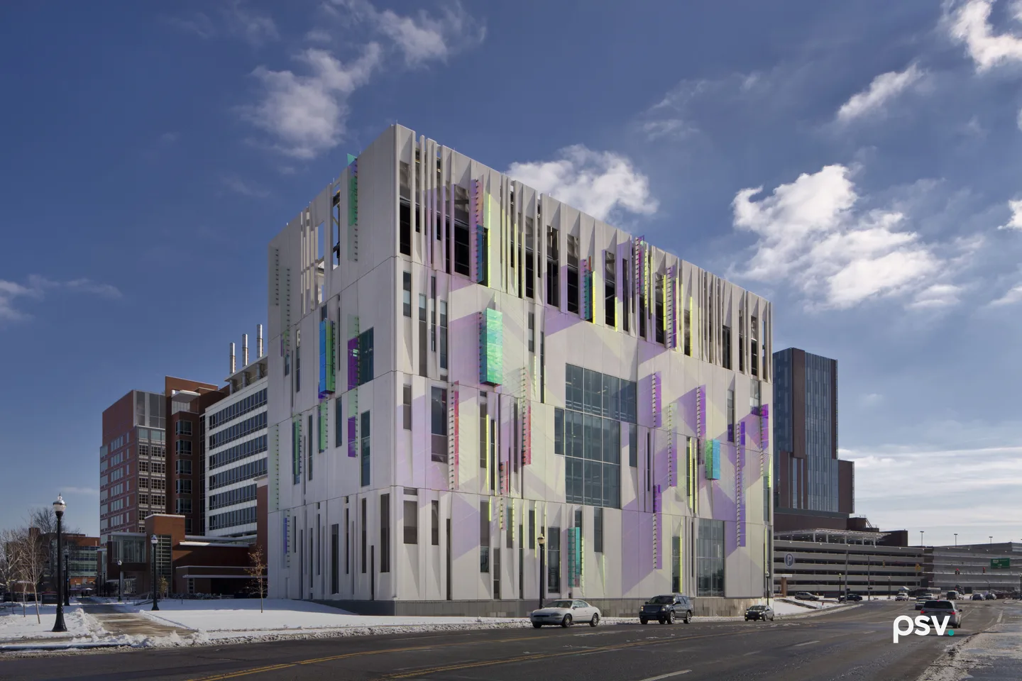 A daylight street-level view of a four-storey central chiller plant on a winter day: a boxy concrete building clad in white panels printed with pale lavender and mint geometric shapes, with narrow vertical fins along the roofline, slotted openings revealing dark mechanical equipment on the top floor, and small iridescent coloured panels scattered across the facade, with a brick hospital block to the left, a parking structure to the right, snow on the verge and a bare asphalt road in front under a blue sky with scattered cloud. A daylight street-level view of a four-storey central chiller plant on a winter day: a boxy concrete building clad in white panels printed with pale lavender and mint geometric shapes, with narrow vertical fins along the roofline, slotted openings revealing dark mechanical equipment on the top floor, and small iridescent coloured panels scattered across the facade, with a brick hospital block to the left, a parking structure to the right, snow on the verge and a bare asphalt road in front under a blue sky with scattered cloud.