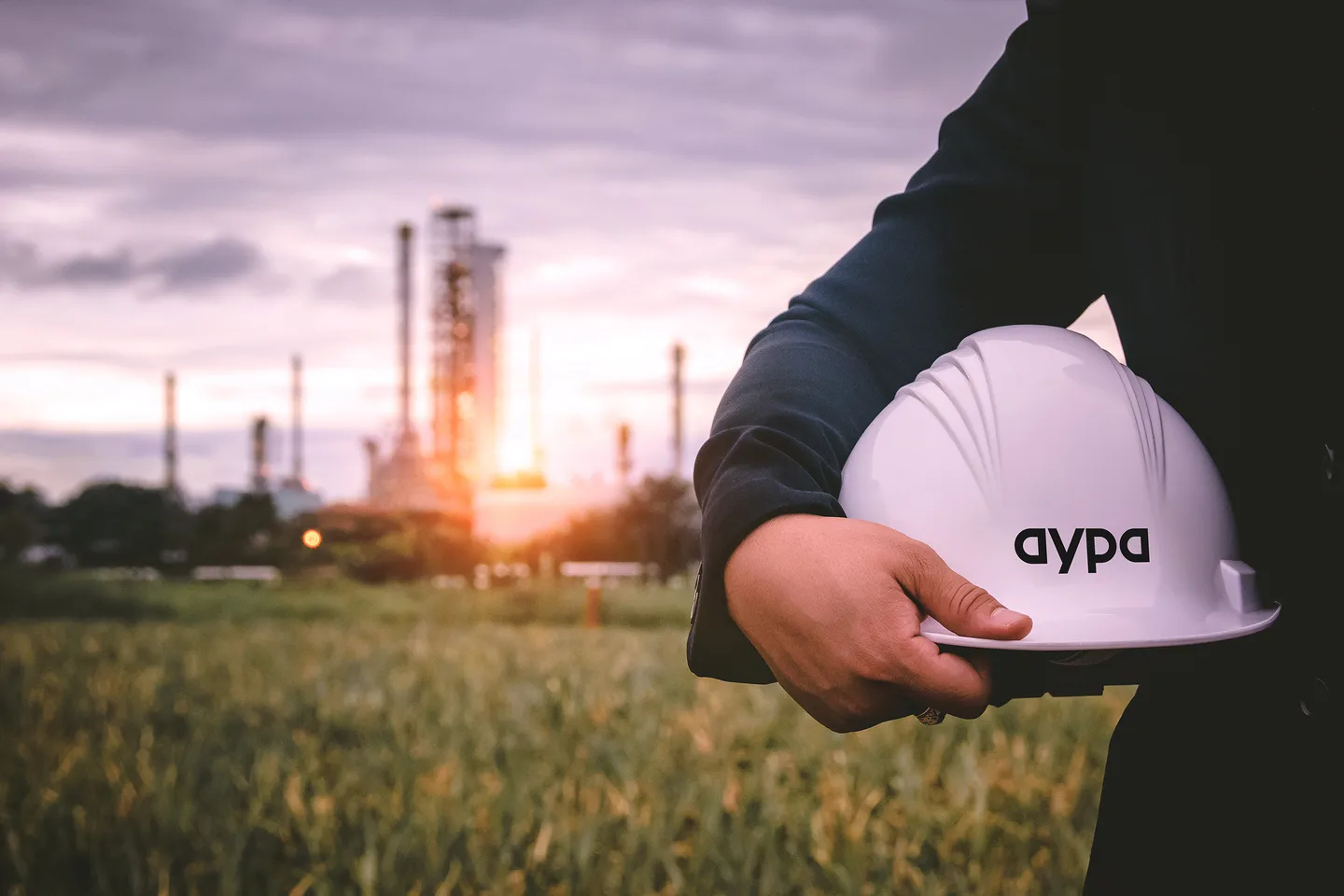 A worker in a dark jacket stands in a grass field at sunset holding a white hard hat marked with the Aypa wordmark, with the stacks and towers of an energy facility silhouetted against the sky behind A worker in a dark jacket stands in a grass field at sunset holding a white hard hat marked with the Aypa wordmark, with the stacks and towers of an energy facility silhouetted against the sky behind
