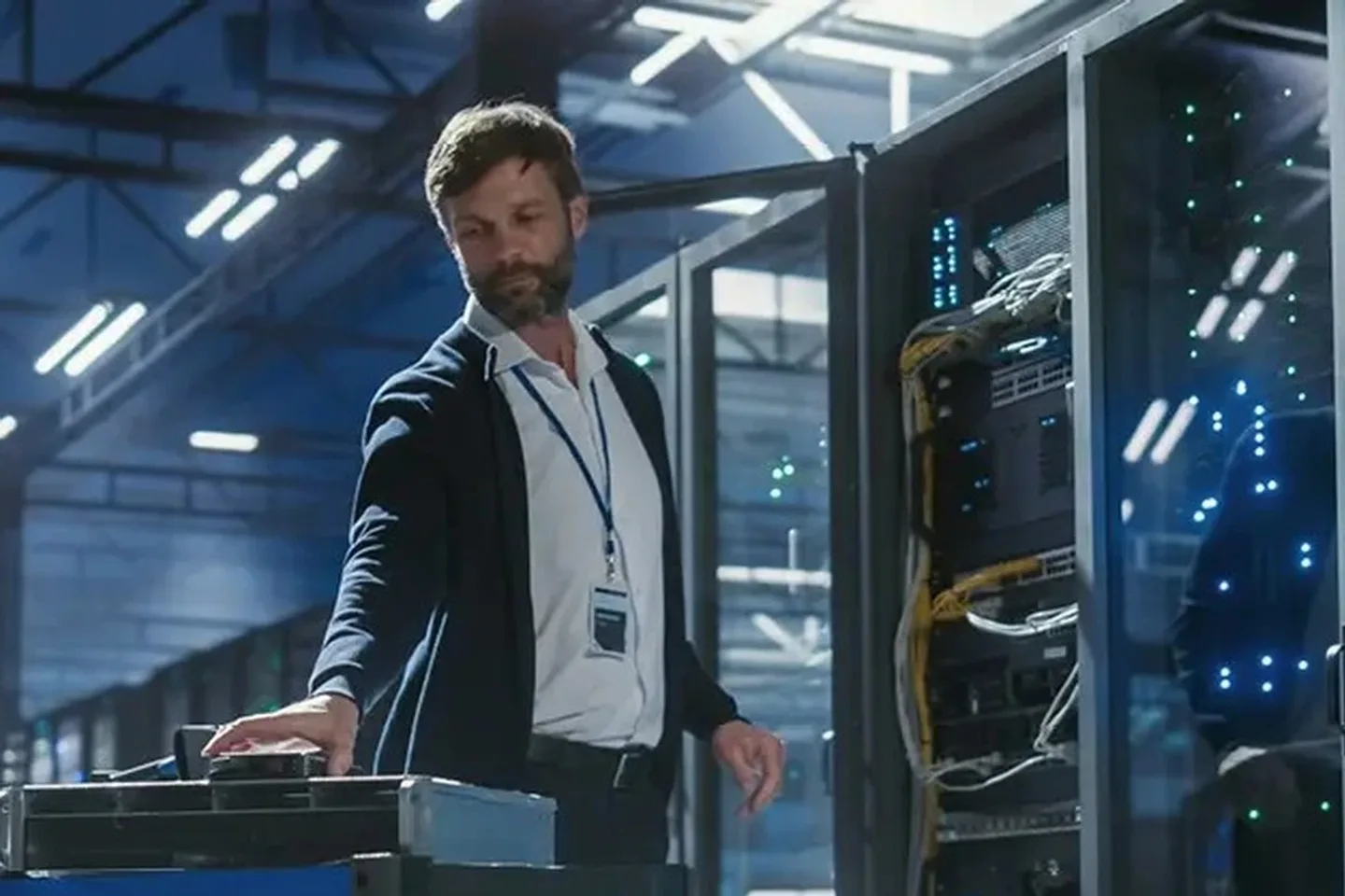 A technician wearing a badge lanyard works at an equipment cart beside an open server cabinet in a brightly lit data hall, cabling and status lights visible in the rack behind him A technician wearing a badge lanyard works at an equipment cart beside an open server cabinet in a brightly lit data hall, cabling and status lights visible in the rack behind him