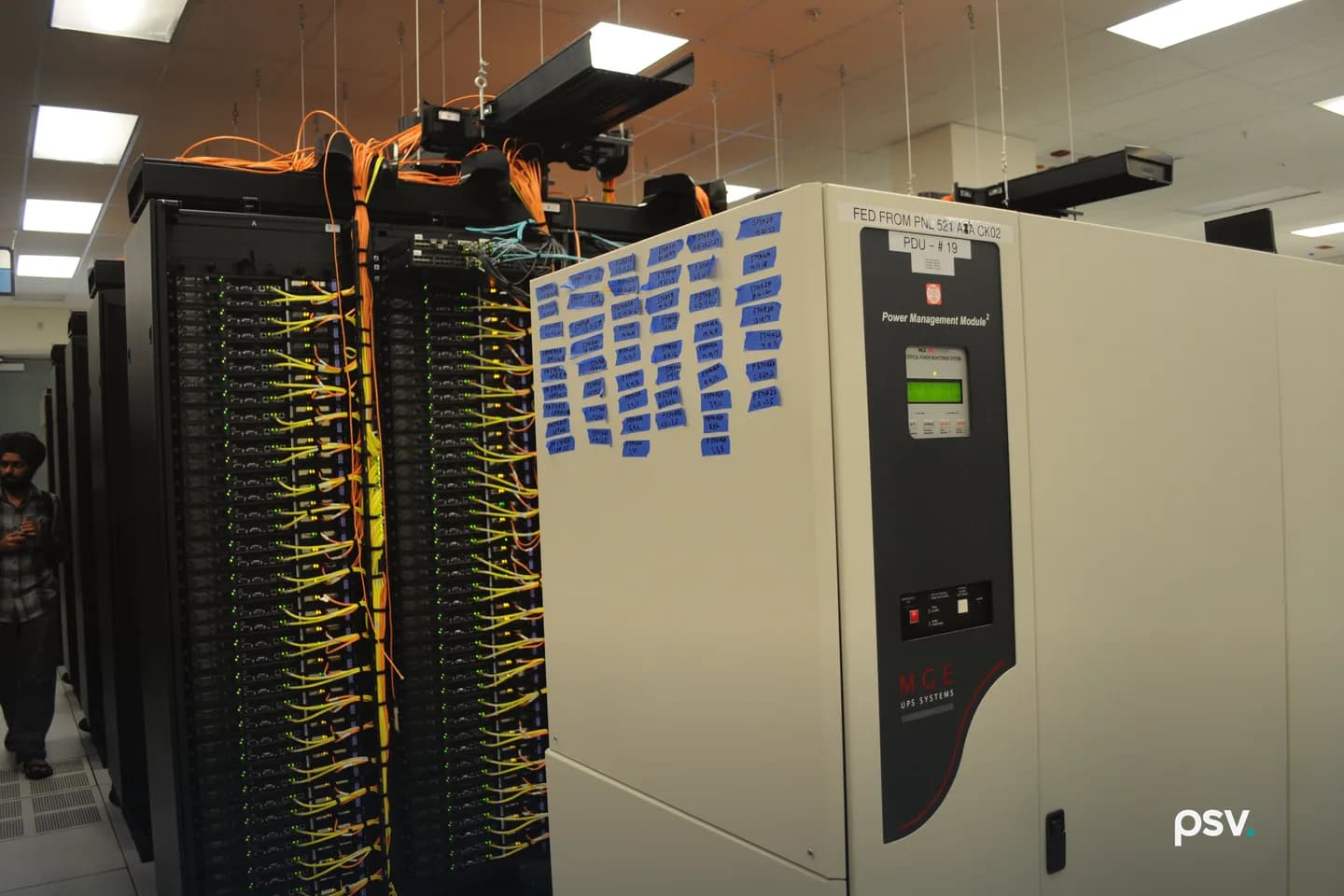 Rows of black server racks in a data hall with bundles of orange and yellow network cables running down their fronts and across overhead trays, beside a tall beige UPS power management cabinet covered in blue tape labels, under a white drop ceiling with fluorescent lights; a person walks in the aisle at far left. Rows of black server racks in a data hall with bundles of orange and yellow network cables running down their fronts and across overhead trays, beside a tall beige UPS power management cabinet covered in blue tape labels, under a white drop ceiling with fluorescent lights; a person walks in the aisle at far left.