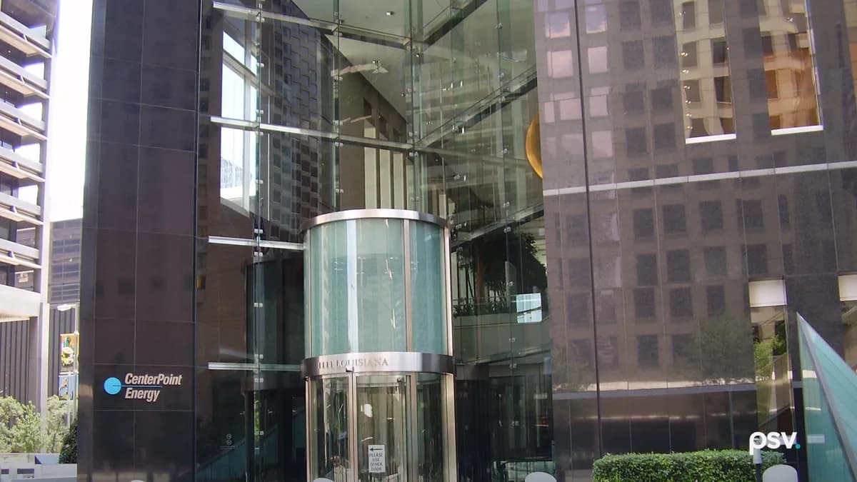 The street-level entrance to a downtown office tower: a cylindrical glass revolving door set into a full-height glass atrium between dark polished granite walls, with a CenterPoint Energy wordmark and logo mounted on the granite at left, planted beds and metal bollards across the paved plaza in front, and neighbouring towers reflected in the glass.
