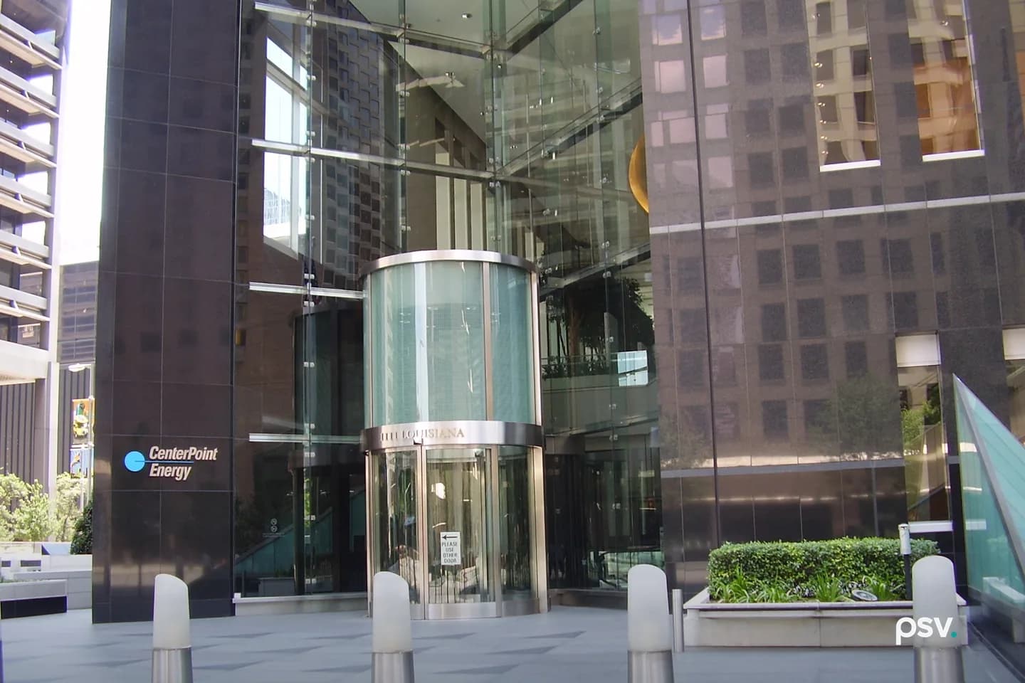 The street-level entrance to a downtown office tower: a cylindrical glass revolving door set into a full-height glass atrium between dark polished granite walls, with a CenterPoint Energy wordmark and logo mounted on the granite at left, planted beds and metal bollards across the paved plaza in front, and neighbouring towers reflected in the glass. The street-level entrance to a downtown office tower: a cylindrical glass revolving door set into a full-height glass atrium between dark polished granite walls, with a CenterPoint Energy wordmark and logo mounted on the granite at left, planted beds and metal bollards across the paved plaza in front, and neighbouring towers reflected in the glass.