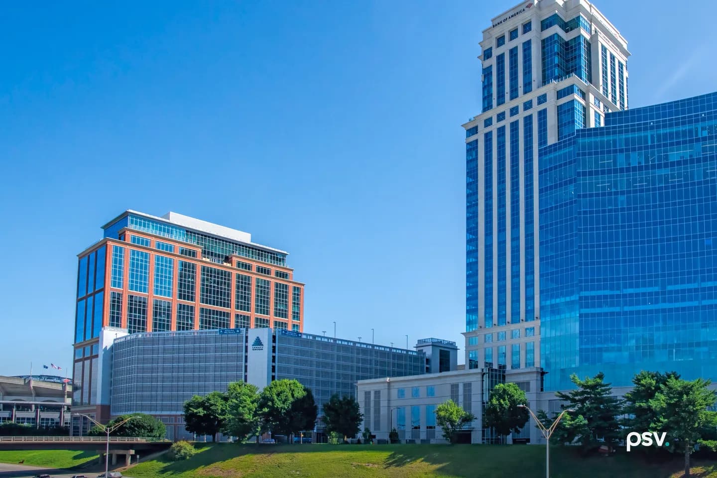 A red-brick and glass office building with a gray parking deck at its base beside a tall white-and-blue glass tower marked Bank of America, with a stadium edge and highway embankment at lower left, under a clear blue sky. A red-brick and glass office building with a gray parking deck at its base beside a tall white-and-blue glass tower marked Bank of America, with a stadium edge and highway embankment at lower left, under a clear blue sky.