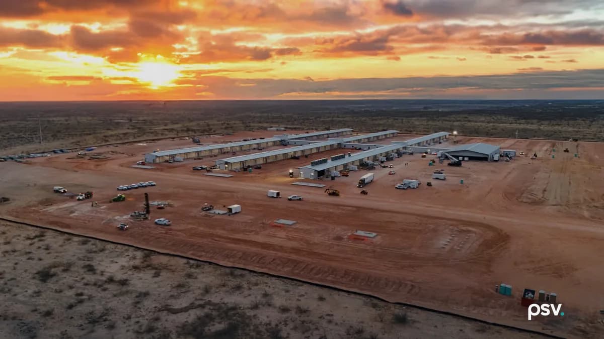 An aerial photograph at sunset of a cleared, graded site of red earth in flat scrubland: four long, low single-storey buildings with pale metal roofs sit in parallel rows at the centre beside a larger dark-roofed shed, with trucks, pickup trucks, trailers and a drilling rig scattered across the dirt in front of them, a dark silt fence curving along the near edge of the cleared ground, and an orange and yellow sky with heavy cloud over a level horizon.