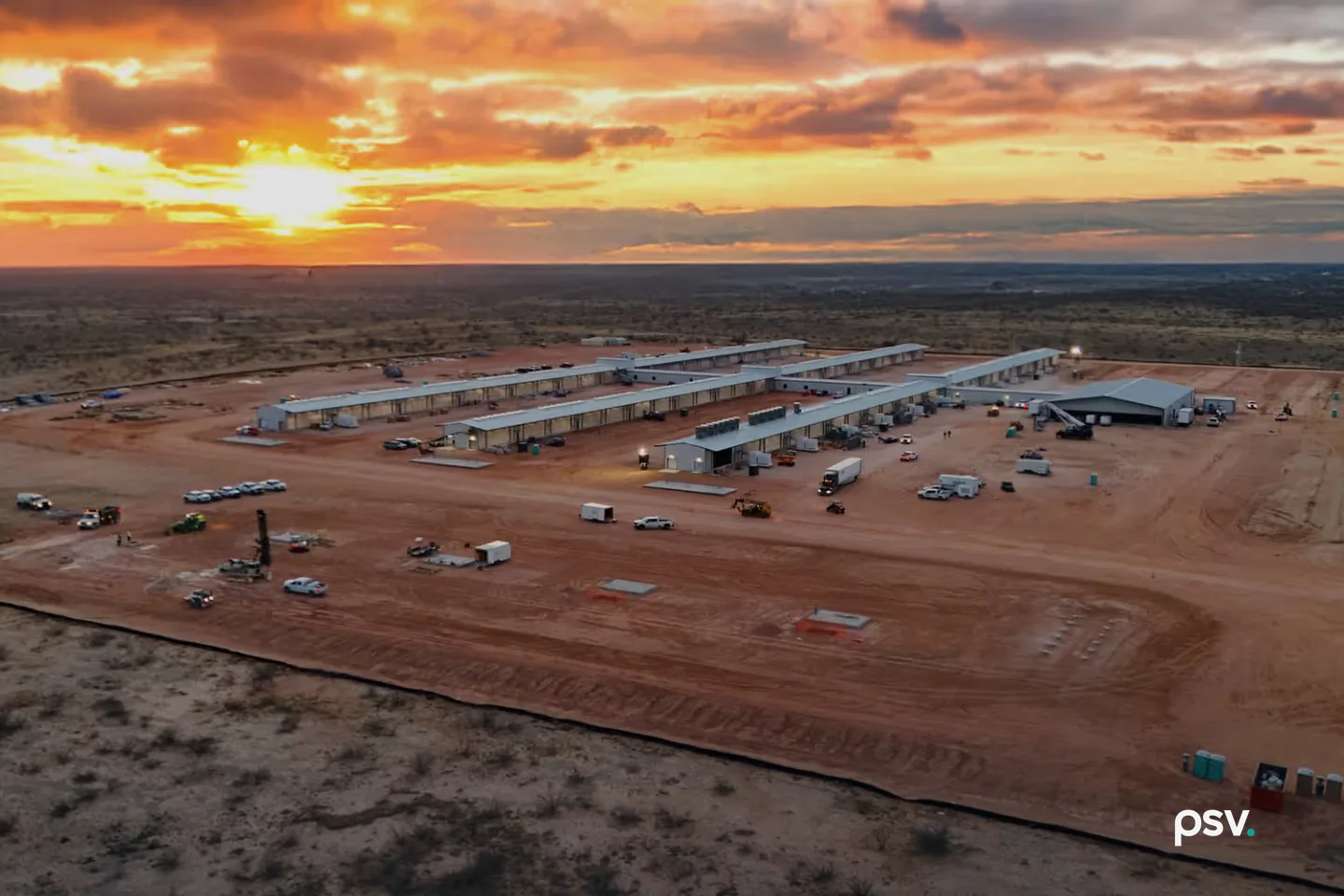 An aerial photograph at sunset of a cleared, graded site of red earth in flat scrubland: four long, low single-storey buildings with pale metal roofs sit in parallel rows at the centre beside a larger dark-roofed shed, with trucks, pickup trucks, trailers and a drilling rig scattered across the dirt in front of them, a dark silt fence curving along the near edge of the cleared ground, and an orange and yellow sky with heavy cloud over a level horizon. An aerial photograph at sunset of a cleared, graded site of red earth in flat scrubland: four long, low single-storey buildings with pale metal roofs sit in parallel rows at the centre beside a larger dark-roofed shed, with trucks, pickup trucks, trailers and a drilling rig scattered across the dirt in front of them, a dark silt fence curving along the near edge of the cleared ground, and an orange and yellow sky with heavy cloud over a level horizon.