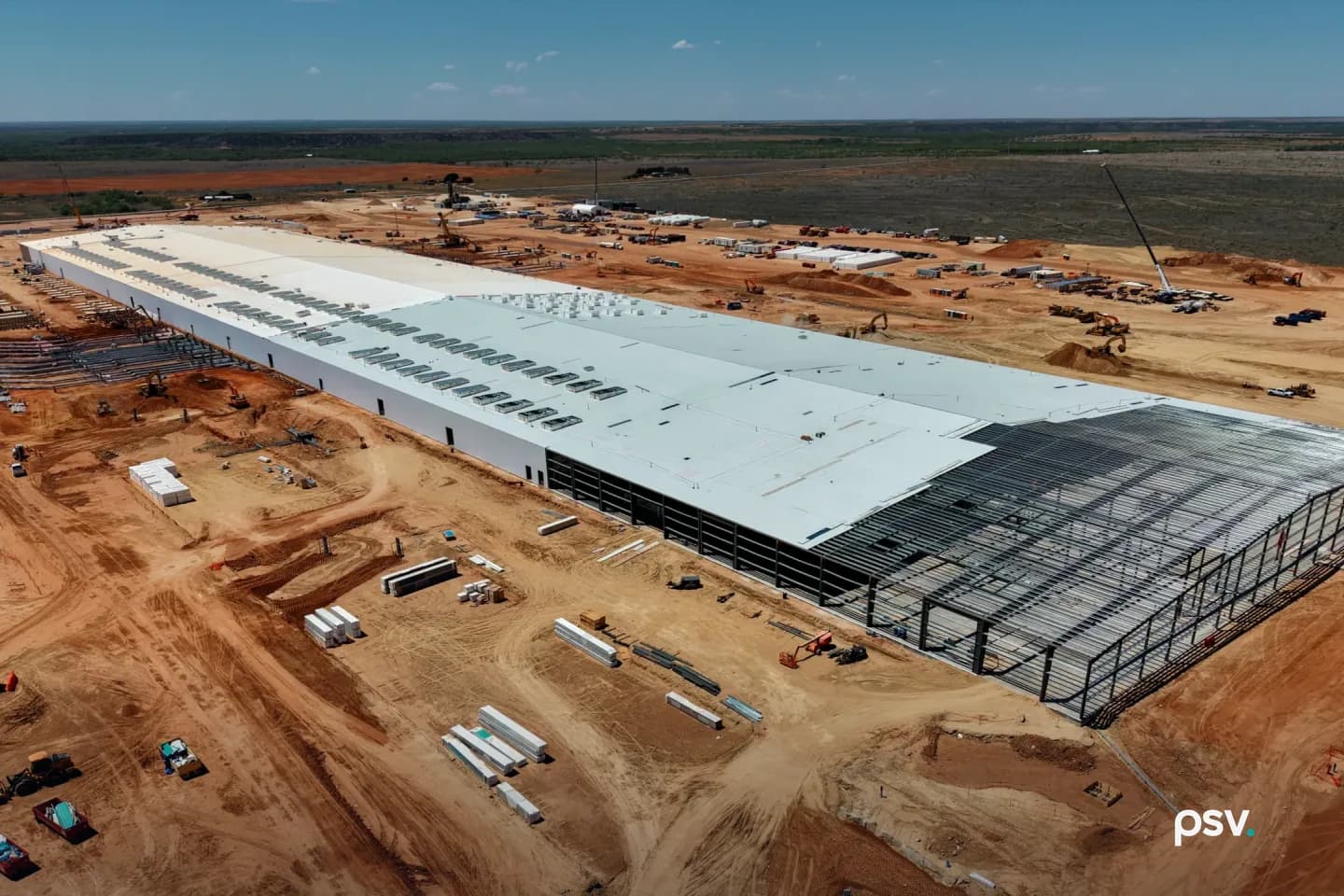 An aerial daylight photograph of a very large single-storey data center hall under construction on graded red earth: a long pale roof lined with two rows of rooftop units runs diagonally from the left edge toward the right, where the building ends in exposed dark steel framing not yet clad, bundles of wall panels and building materials lie on the dirt in the foreground with small vehicles at the lower left, excavators, soil piles and a tall crane work across the site behind it, and flat green-grey scrubland stretches to the horizon under a blue sky with a few small clouds. An aerial daylight photograph of a very large single-storey data center hall under construction on graded red earth: a long pale roof lined with two rows of rooftop units runs diagonally from the left edge toward the right, where the building ends in exposed dark steel framing not yet clad, bundles of wall panels and building materials lie on the dirt in the foreground with small vehicles at the lower left, excavators, soil piles and a tall crane work across the site behind it, and flat green-grey scrubland stretches to the horizon under a blue sky with a few small clouds.