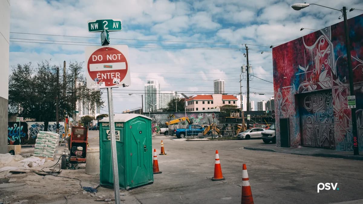 A daylight street corner in Miami’s Wynwood neighborhood under a partly cloudy sky, with a NW 2 Av street sign above a Do Not Enter sign in the foreground, a green portable toilet, cement bags and a generator on a dusty construction lot, orange traffic cones in the road, a warehouse wall covered in a red and blue mural at the right, an excavator and pickup trucks behind a graffiti-covered wall, and downtown Miami high-rise towers in the background.