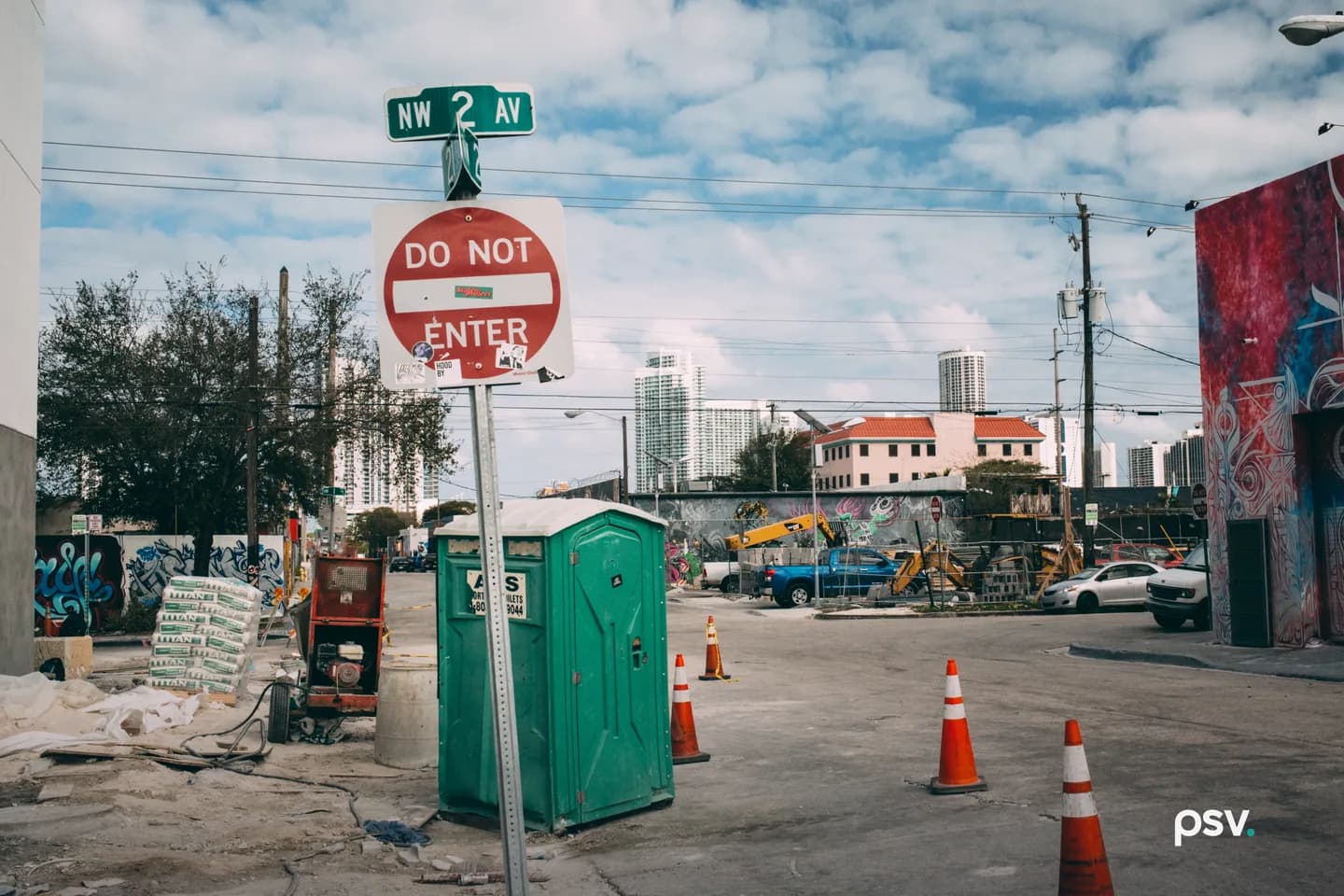 A daylight street corner in Miami’s Wynwood neighborhood under a partly cloudy sky, with a NW 2 Av street sign above a Do Not Enter sign in the foreground, a green portable toilet, cement bags and a generator on a dusty construction lot, orange traffic cones in the road, a warehouse wall covered in a red and blue mural at the right, an excavator and pickup trucks behind a graffiti-covered wall, and downtown Miami high-rise towers in the background. A daylight street corner in Miami’s Wynwood neighborhood under a partly cloudy sky, with a NW 2 Av street sign above a Do Not Enter sign in the foreground, a green portable toilet, cement bags and a generator on a dusty construction lot, orange traffic cones in the road, a warehouse wall covered in a red and blue mural at the right, an excavator and pickup trucks behind a graffiti-covered wall, and downtown Miami high-rise towers in the background.