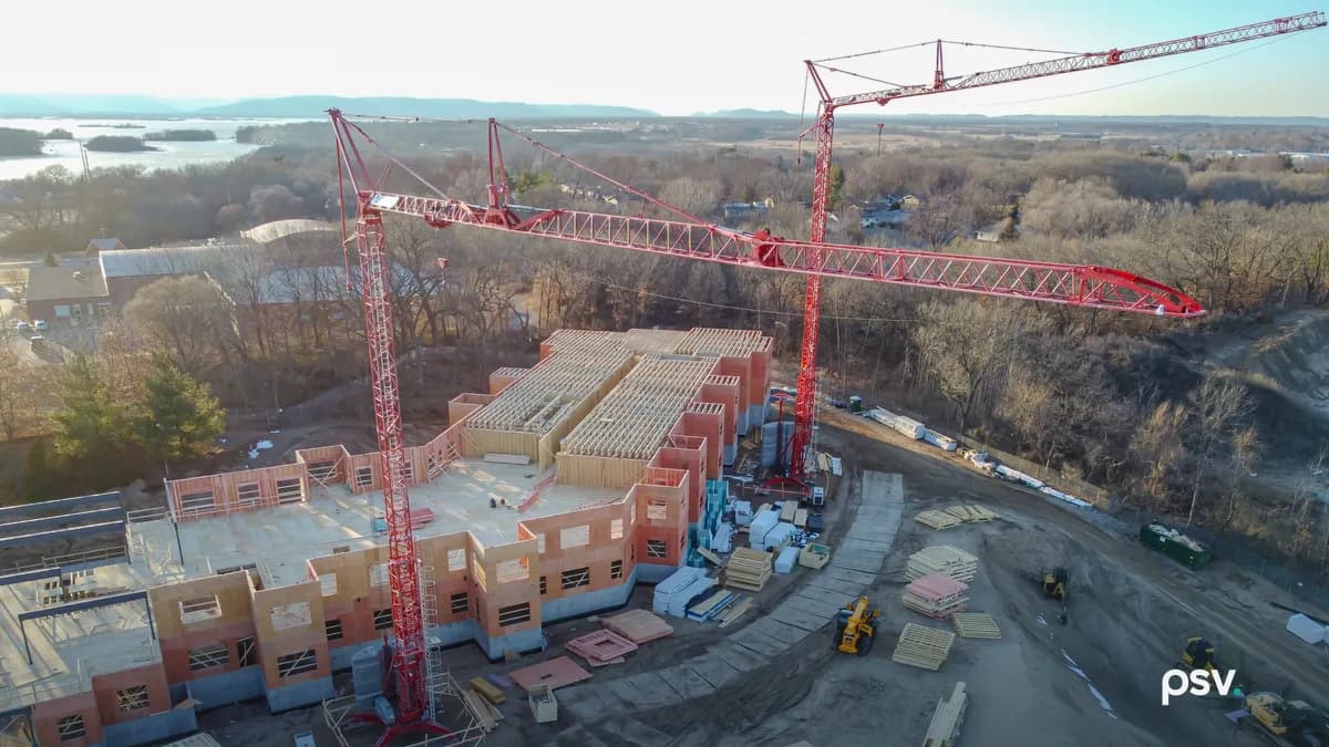 Aerial view of a mid-rise building under construction: two red tower cranes standing over a partly framed structure of prefabricated wood wall panels on a concrete podium, open floor joists across the top level, stacks of panels, lumber and insulation laid out on the graded dirt around it, small yellow loaders parked on the haul road, with bare winter trees and a river beyond