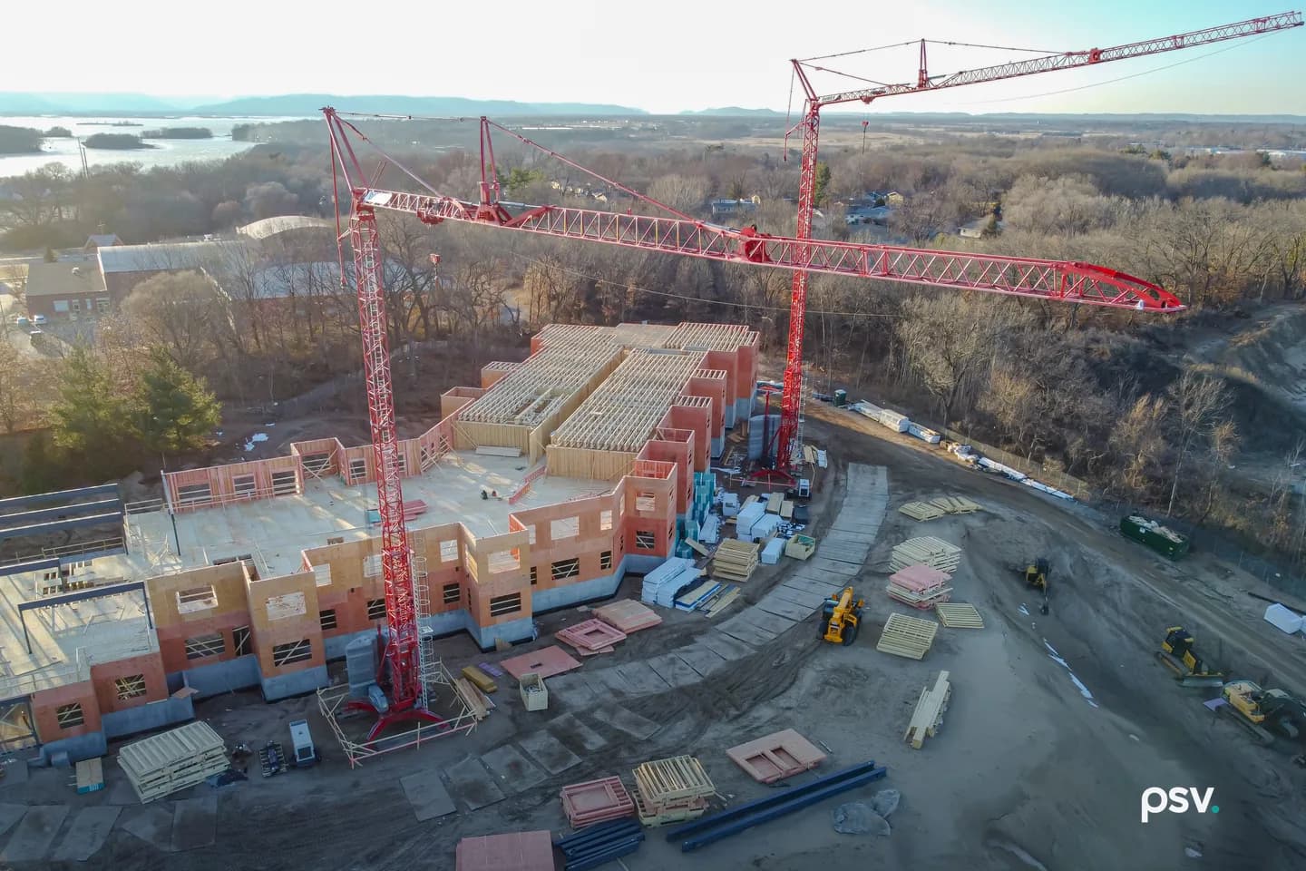 Aerial view of a mid-rise building under construction: two red tower cranes standing over a partly framed structure of prefabricated wood wall panels on a concrete podium, open floor joists across the top level, stacks of panels, lumber and insulation laid out on the graded dirt around it, small yellow loaders parked on the haul road, with bare winter trees and a river beyond Aerial view of a mid-rise building under construction: two red tower cranes standing over a partly framed structure of prefabricated wood wall panels on a concrete podium, open floor joists across the top level, stacks of panels, lumber and insulation laid out on the graded dirt around it, small yellow loaders parked on the haul road, with bare winter trees and a river beyond