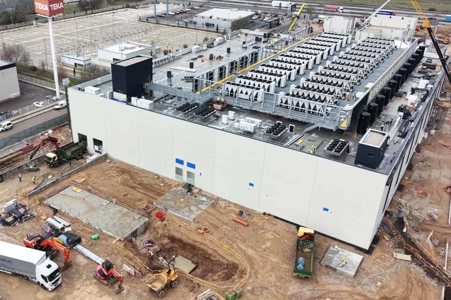 Aerial view of a Coravel data hall under construction: rows of rooftop chiller units being installed on the long white building, cranes and construction crews working the graded site below Aerial view of a Coravel data hall under construction: rows of rooftop chiller units being installed on the long white building, cranes and construction crews working the graded site below