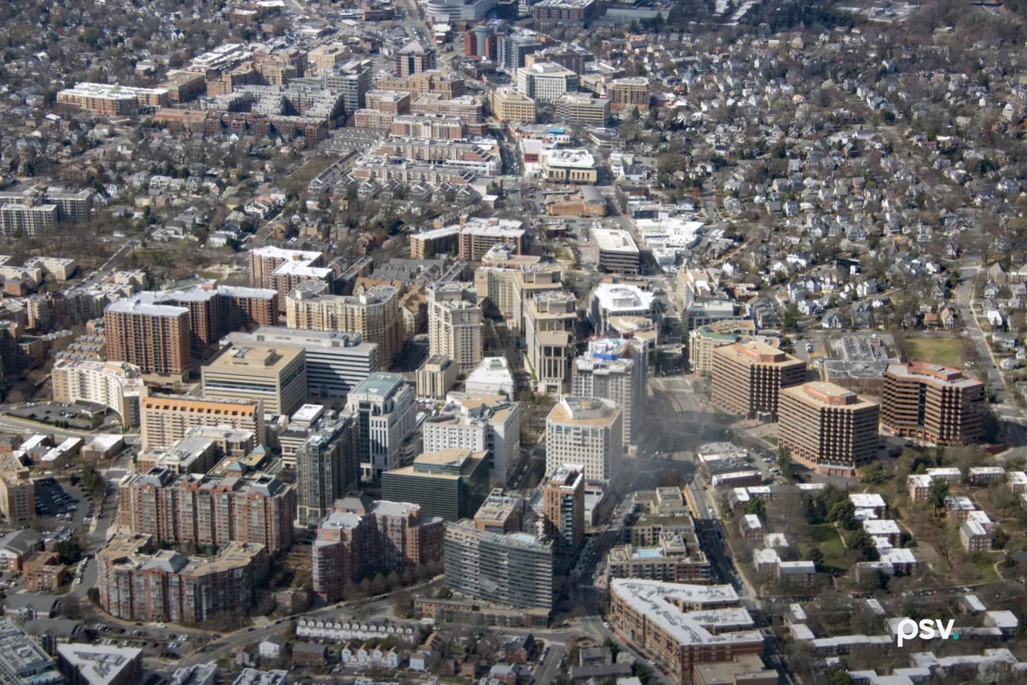 An oblique aerial view of a dense Arlington County, Virginia district in late winter: a corridor of mid-rise brick and concrete office and apartment buildings with flat rooftops runs through the center, bordered by blocks of single-family houses and bare trees, with a faint haze of smoke drifting across one street. An oblique aerial view of a dense Arlington County, Virginia district in late winter: a corridor of mid-rise brick and concrete office and apartment buildings with flat rooftops runs through the center, bordered by blocks of single-family houses and bare trees, with a faint haze of smoke drifting across one street.