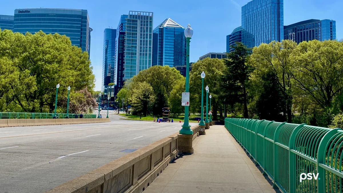 A daylight view along the Key Bridge sidewalk toward the Rosslyn skyline in Arlington, Virginia: a row of green cast-iron lampposts and a green metal railing running into the distance beside an empty multi-lane roadway, leafy trees on both sides, and a cluster of modern glass and stone office towers rising behind them under a clear blue sky.