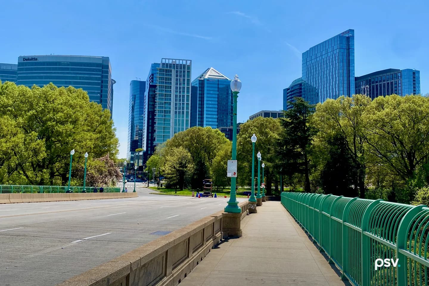 A daylight view along the Key Bridge sidewalk toward the Rosslyn skyline in Arlington, Virginia: a row of green cast-iron lampposts and a green metal railing running into the distance beside an empty multi-lane roadway, leafy trees on both sides, and a cluster of modern glass and stone office towers rising behind them under a clear blue sky. A daylight view along the Key Bridge sidewalk toward the Rosslyn skyline in Arlington, Virginia: a row of green cast-iron lampposts and a green metal railing running into the distance beside an empty multi-lane roadway, leafy trees on both sides, and a cluster of modern glass and stone office towers rising behind them under a clear blue sky.