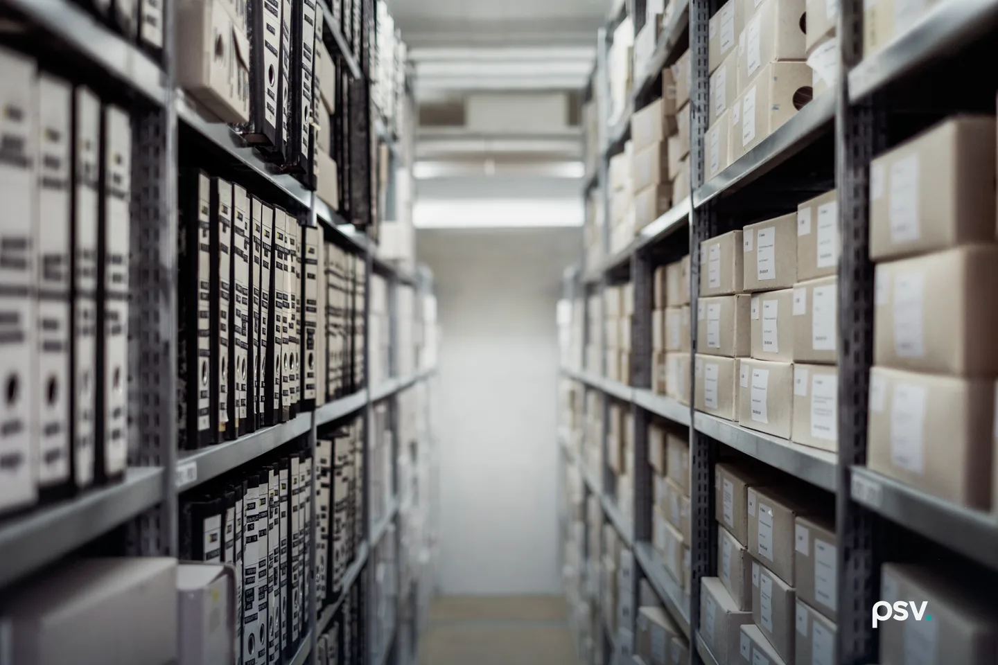 A narrow aisle between tall metal shelving in a document archive, both sides stacked with labeled cardboard storage boxes and ring binders that recede into soft focus toward a lit wall at the end. A narrow aisle between tall metal shelving in a document archive, both sides stacked with labeled cardboard storage boxes and ring binders that recede into soft focus toward a lit wall at the end.
