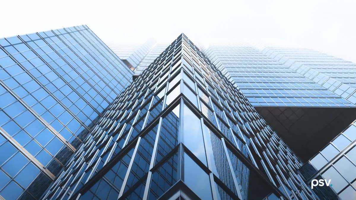A low upward view of a blue glass office tower whose mirrored curtain wall meets at a sharp vertical corner, with a lower glazed wing extending to the left against a bright overcast sky.