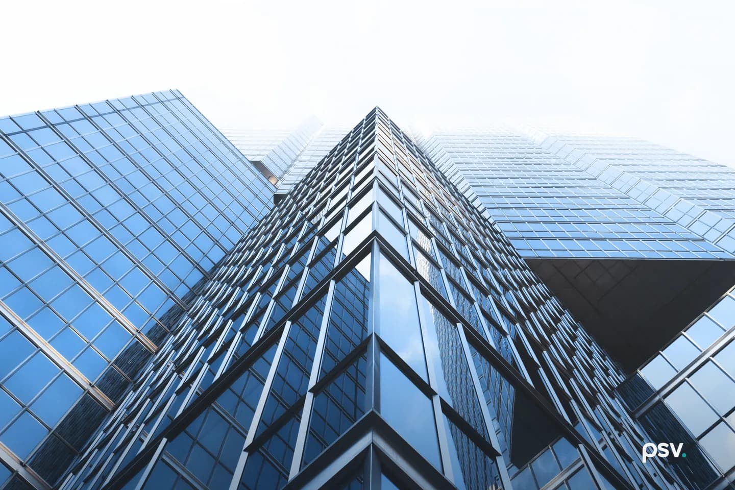 A low upward view of a blue glass office tower whose mirrored curtain wall meets at a sharp vertical corner, with a lower glazed wing extending to the left against a bright overcast sky. A low upward view of a blue glass office tower whose mirrored curtain wall meets at a sharp vertical corner, with a lower glazed wing extending to the left against a bright overcast sky.