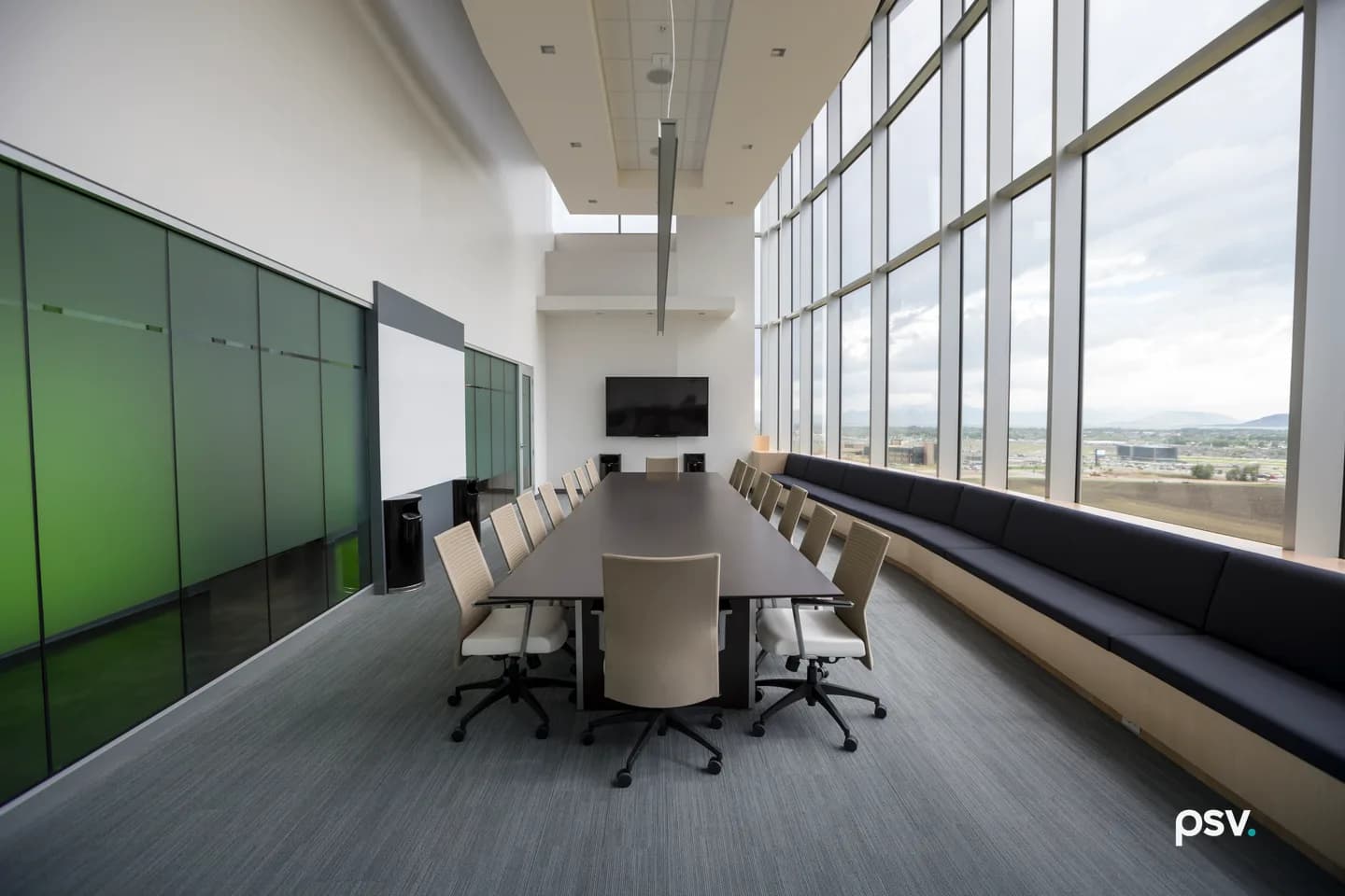 An empty double-height boardroom in an office building: a long charcoal table ringed by taupe ribbed-back chairs with white leather seats, set beside a faceted floor-to-ceiling glass wall and a long dark padded bench that curves along the windows, with green glass panels filling the left wall and a switched-off flat screen on the far wall. An empty double-height boardroom in an office building: a long charcoal table ringed by taupe ribbed-back chairs with white leather seats, set beside a faceted floor-to-ceiling glass wall and a long dark padded bench that curves along the windows, with green glass panels filling the left wall and a switched-off flat screen on the far wall.