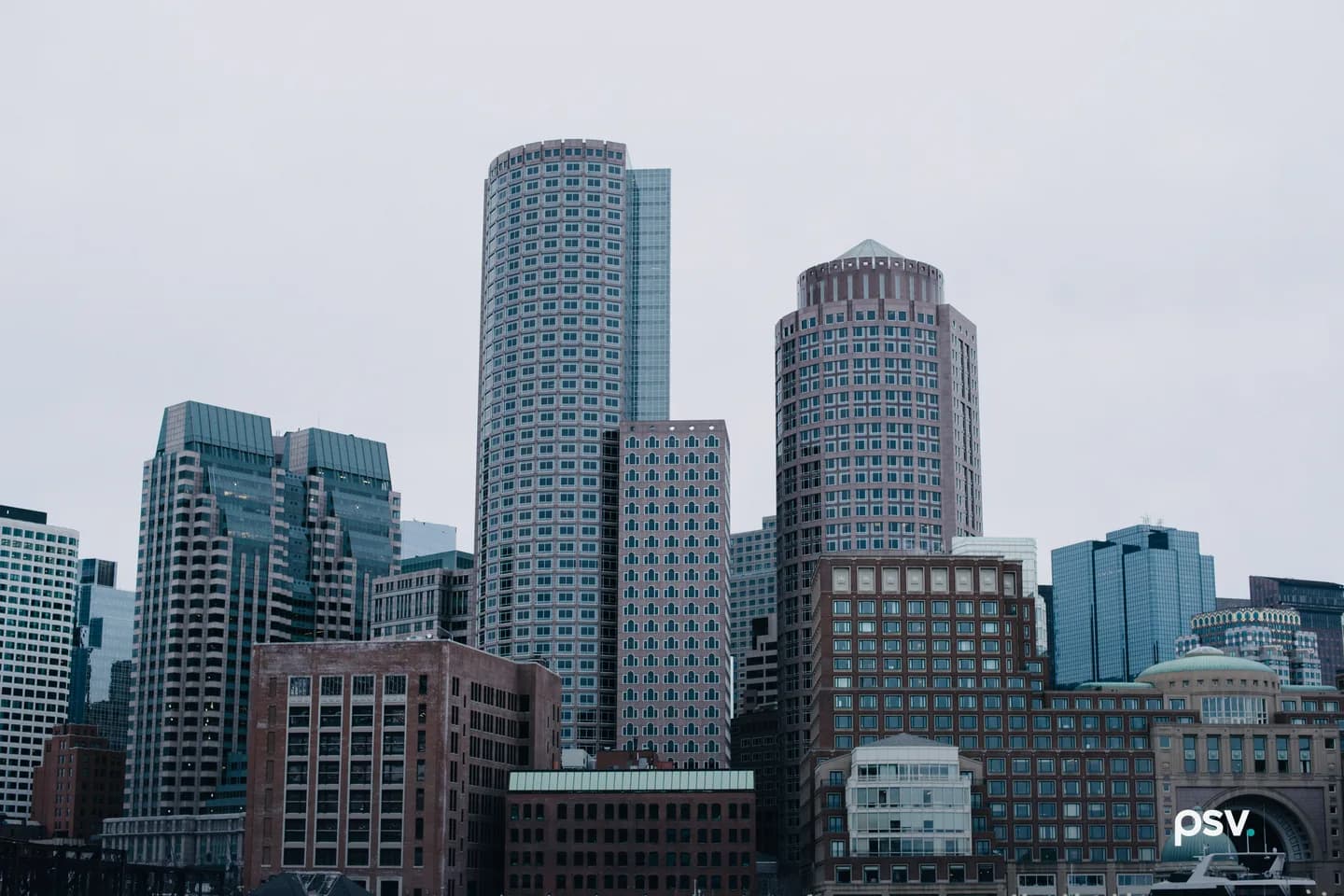 Dense cluster of downtown Boston high-rise office buildings under a flat gray sky, with a precast punched-window tower at center and a glass slab behind it, and brick and concrete mid-rise blocks in front. Dense cluster of downtown Boston high-rise office buildings under a flat gray sky, with a precast punched-window tower at center and a glass slab behind it, and brick and concrete mid-rise blocks in front.