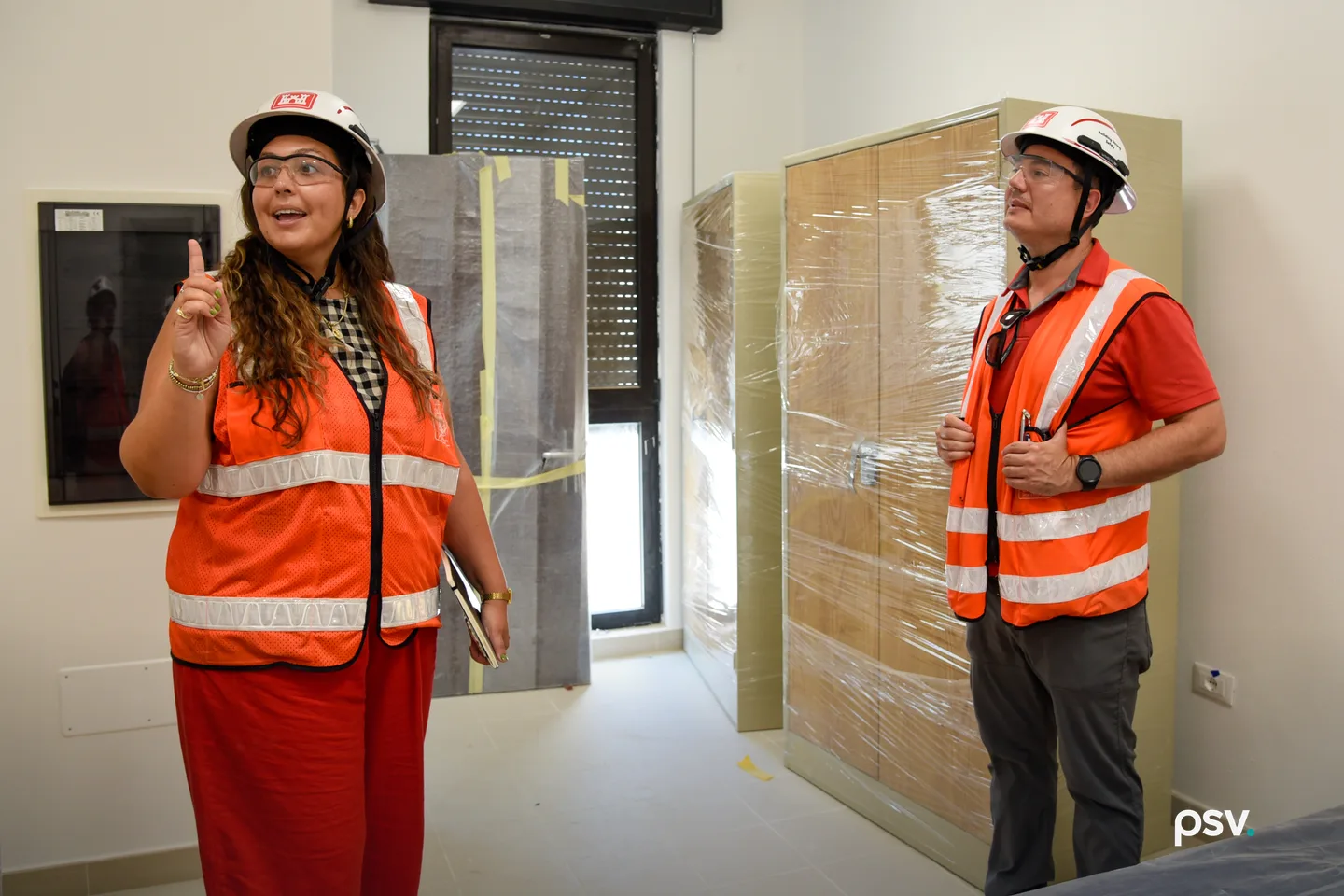 Two building inspectors in orange safety vests, white hard hats and safety glasses stand in a newly renovated interior room. The woman at left holds a notebook and points upward at overhead work while the man at right listens, beside a wall panel and plastic-wrapped cabinetry. Two building inspectors in orange safety vests, white hard hats and safety glasses stand in a newly renovated interior room. The woman at left holds a notebook and points upward at overhead work while the man at right listens, beside a wall panel and plastic-wrapped cabinetry.