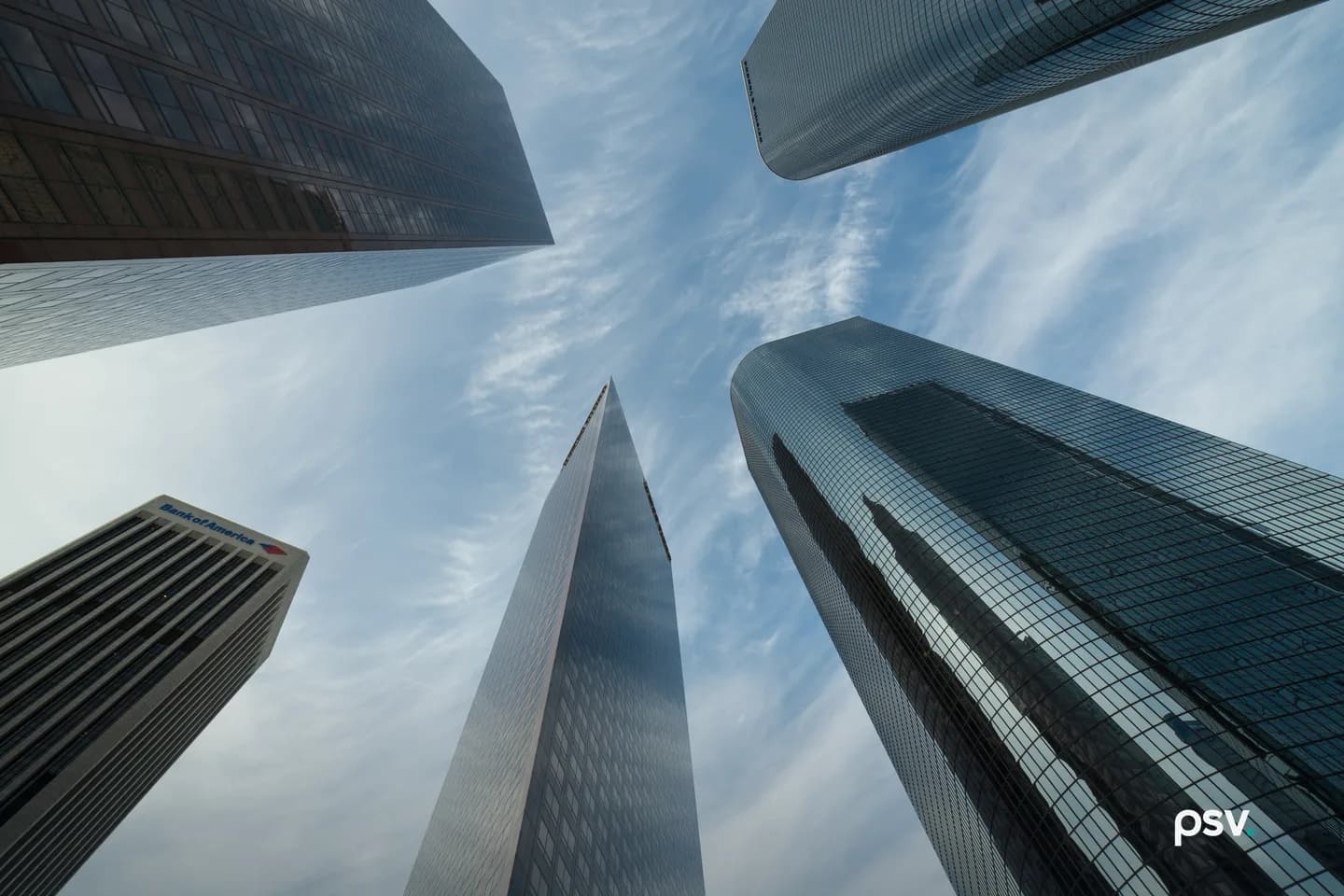 Upward view from street level of five glass and stone skyscrapers in downtown Los Angeles converging toward a blue sky with thin cirrus clouds, their facades reflecting daylight. Upward view from street level of five glass and stone skyscrapers in downtown Los Angeles converging toward a blue sky with thin cirrus clouds, their facades reflecting daylight.
