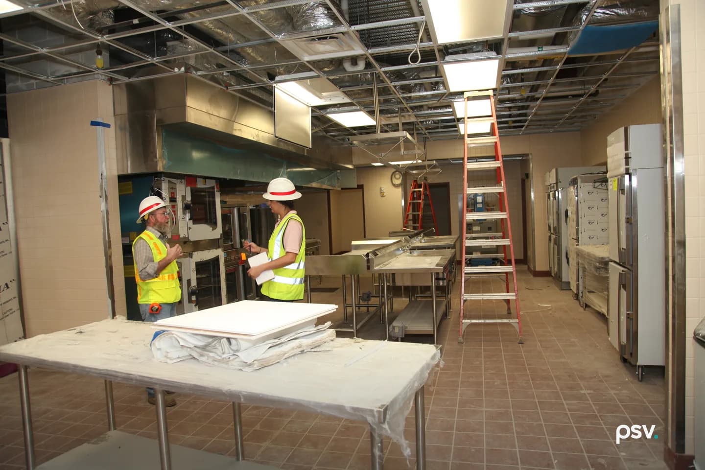 A bearded man in a hard hat and lime-yellow hi-vis vest with orange banding talks with a woman in a hard hat holding a notepad and pen, inside a nearly finished commercial kitchen with stainless prep tables, ovens and exposed ceiling ductwork. A bearded man in a hard hat and lime-yellow hi-vis vest with orange banding talks with a woman in a hard hat holding a notepad and pen, inside a nearly finished commercial kitchen with stainless prep tables, ovens and exposed ceiling ductwork.