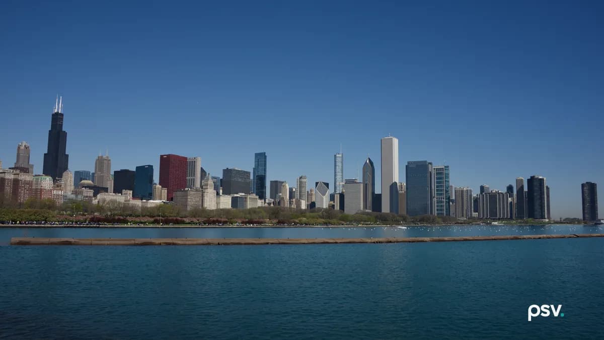 Daylight view across open blue water toward a dense downtown skyline of office and residential towers, with a low stone breakwater in the foreground, a green park strip along the shoreline and a clear sky above.
