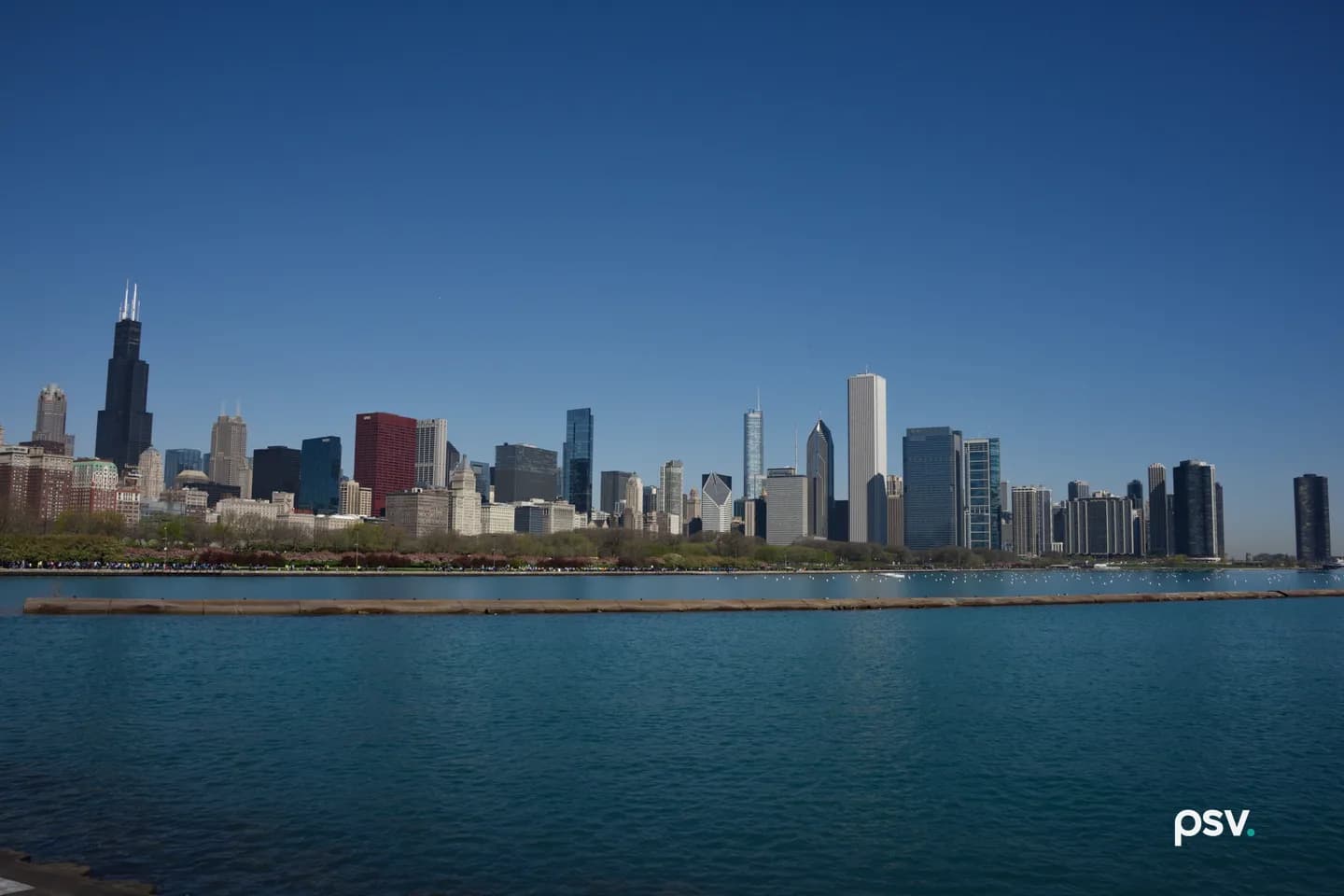 Daylight view across open blue water toward a dense downtown skyline of office and residential towers, with a low stone breakwater in the foreground, a green park strip along the shoreline and a clear sky above. Daylight view across open blue water toward a dense downtown skyline of office and residential towers, with a low stone breakwater in the foreground, a green park strip along the shoreline and a clear sky above.