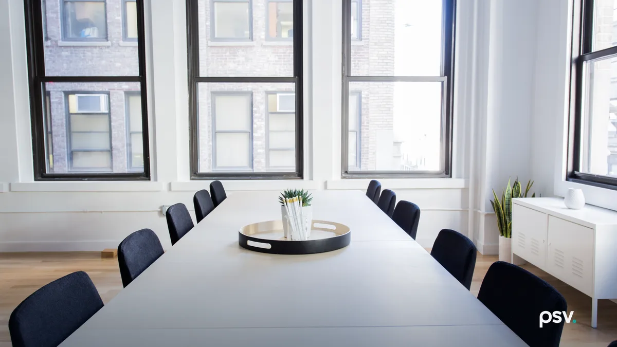 An empty conference room with a long white table, a row of dark chairs down each side, a black tray holding a cup of pens, three tall black framed windows looking onto a brick building across the street, a plant and a low white cabinet.